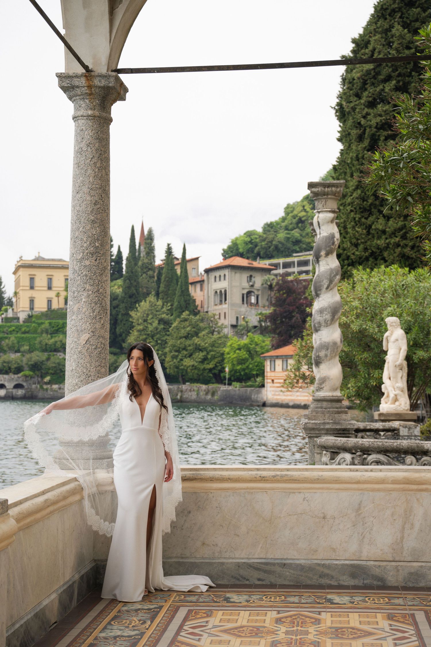 Bride in white gown with veil on marble terrace overlooking Lake Como, Italy, with historic villa in background.
