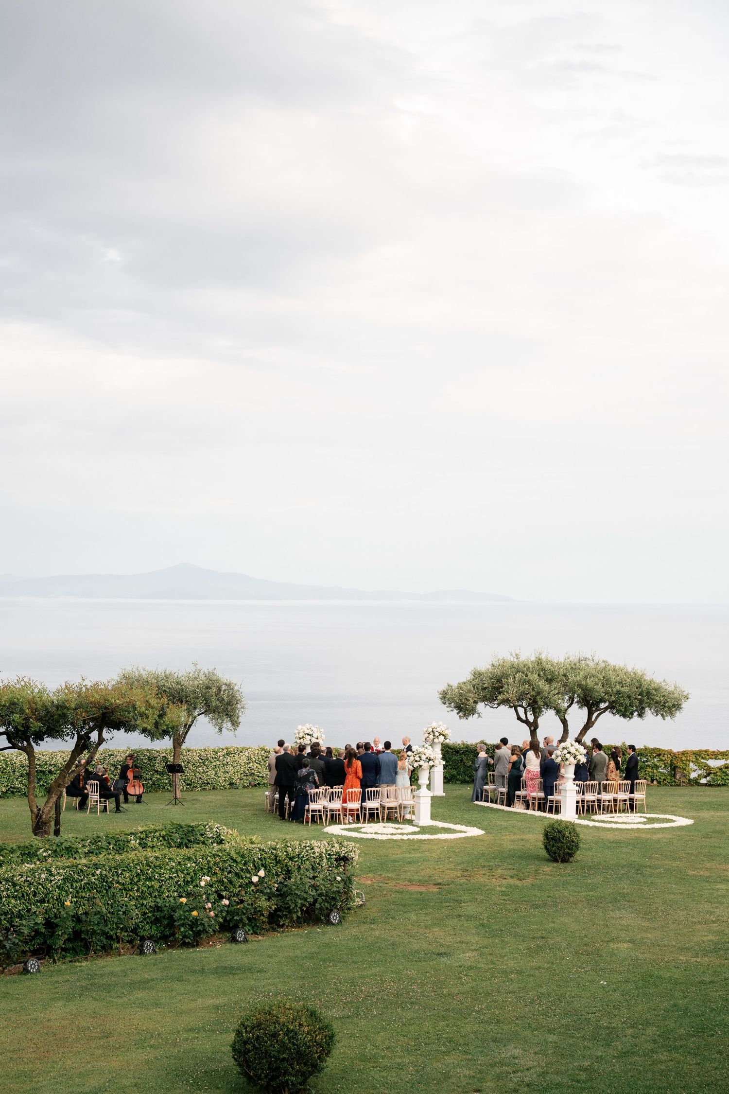 Outdoor wedding ceremony on lush green lawn overlooking the sea with guests seated between olive trees.