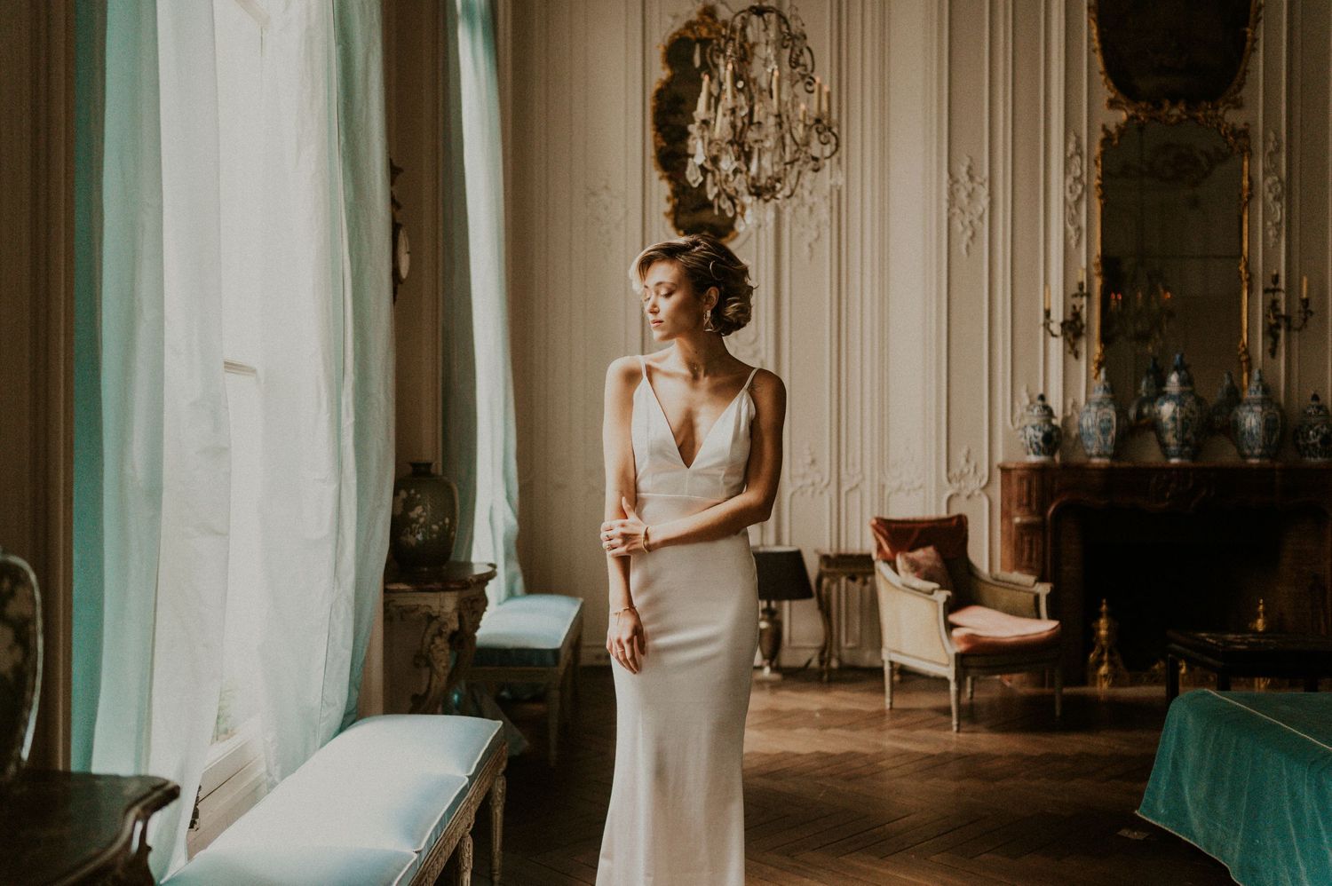 Elegant bride in fitted dress stands by window in ornate Parisian room with crystal chandelier.