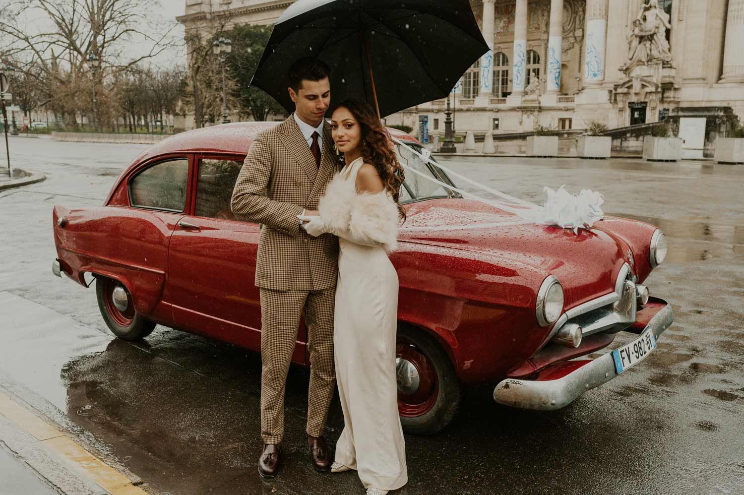 A couple stands with a black umbrella next to a vintage red car on a rainy day.