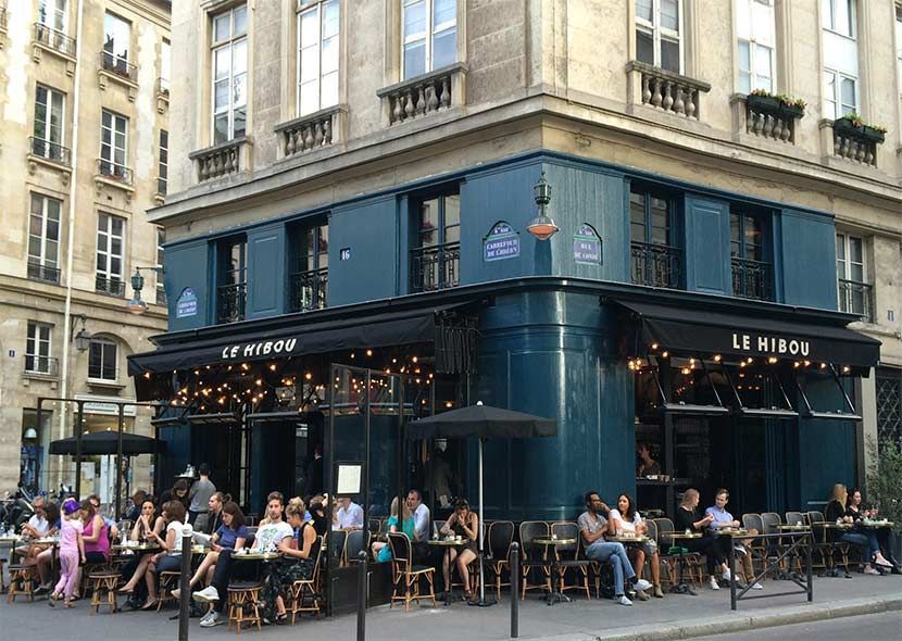 Busy corner cafe in Paris with blue exterior and outdoor seating filled with patrons.