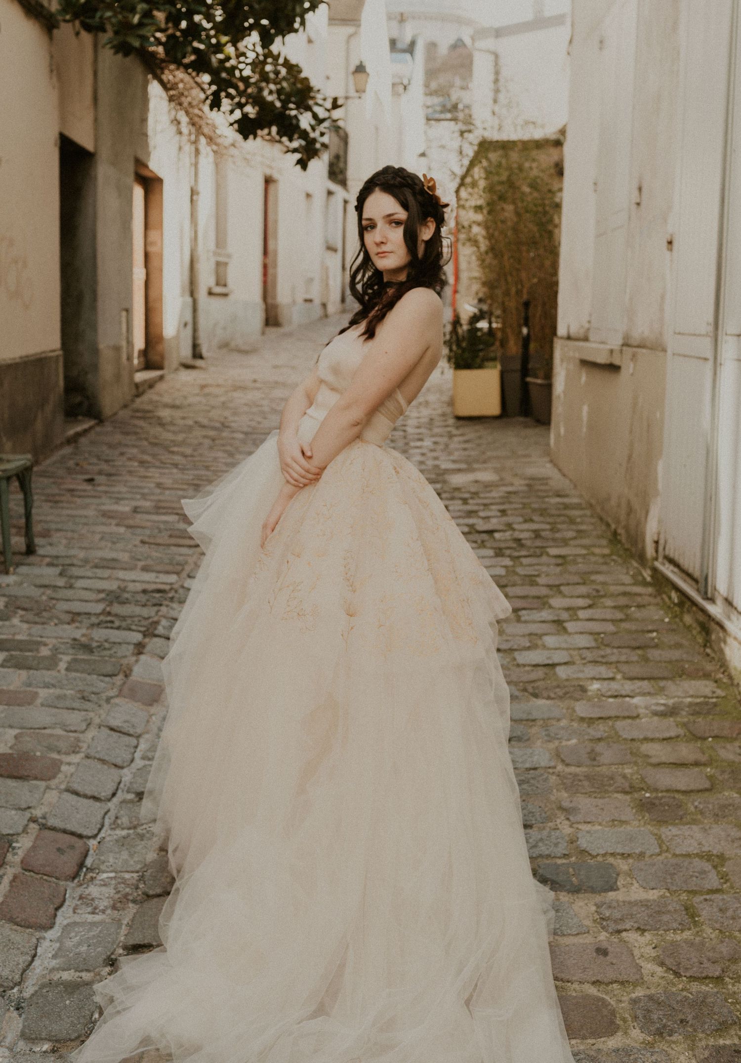 A bride in a champagne-colored ballgown poses elegantly against white walls on a historic street.