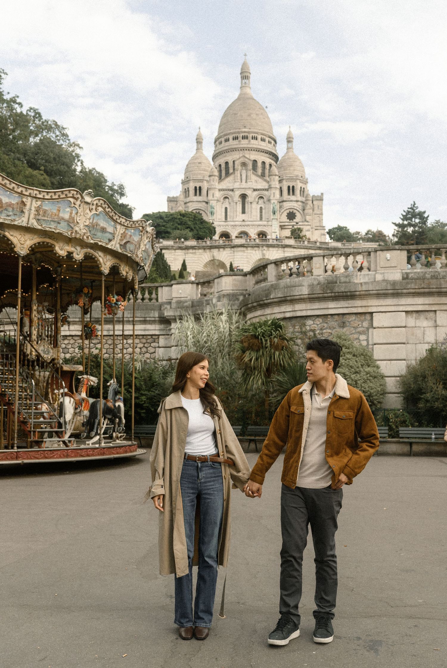 A couple holds hands near a carousel with the iconic Sacré-Cœur Basilica in Paris behind them.