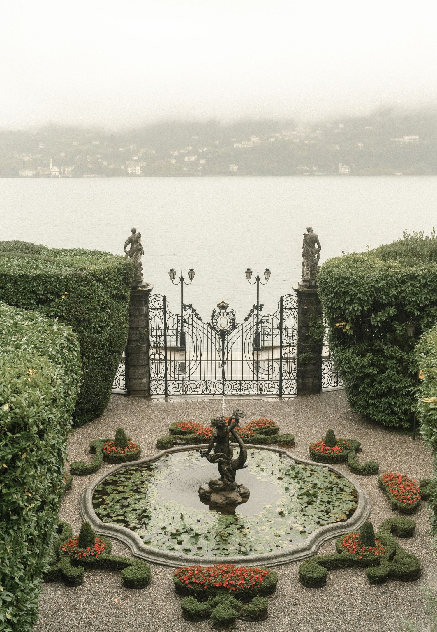 Ornate garden gate with fountain and manicured hedges overlooking a misty lake landscape.