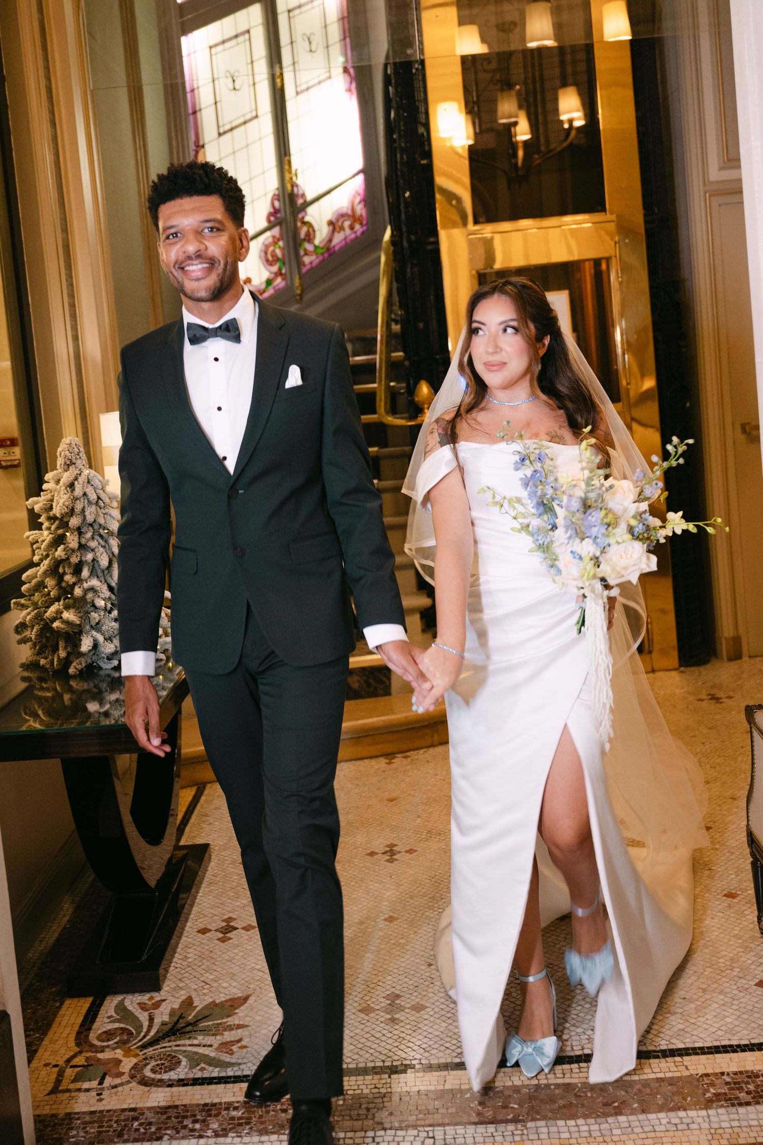 Elegant bride and groom walking hand in hand through ornate hotel lobby in Paris wedding attire.