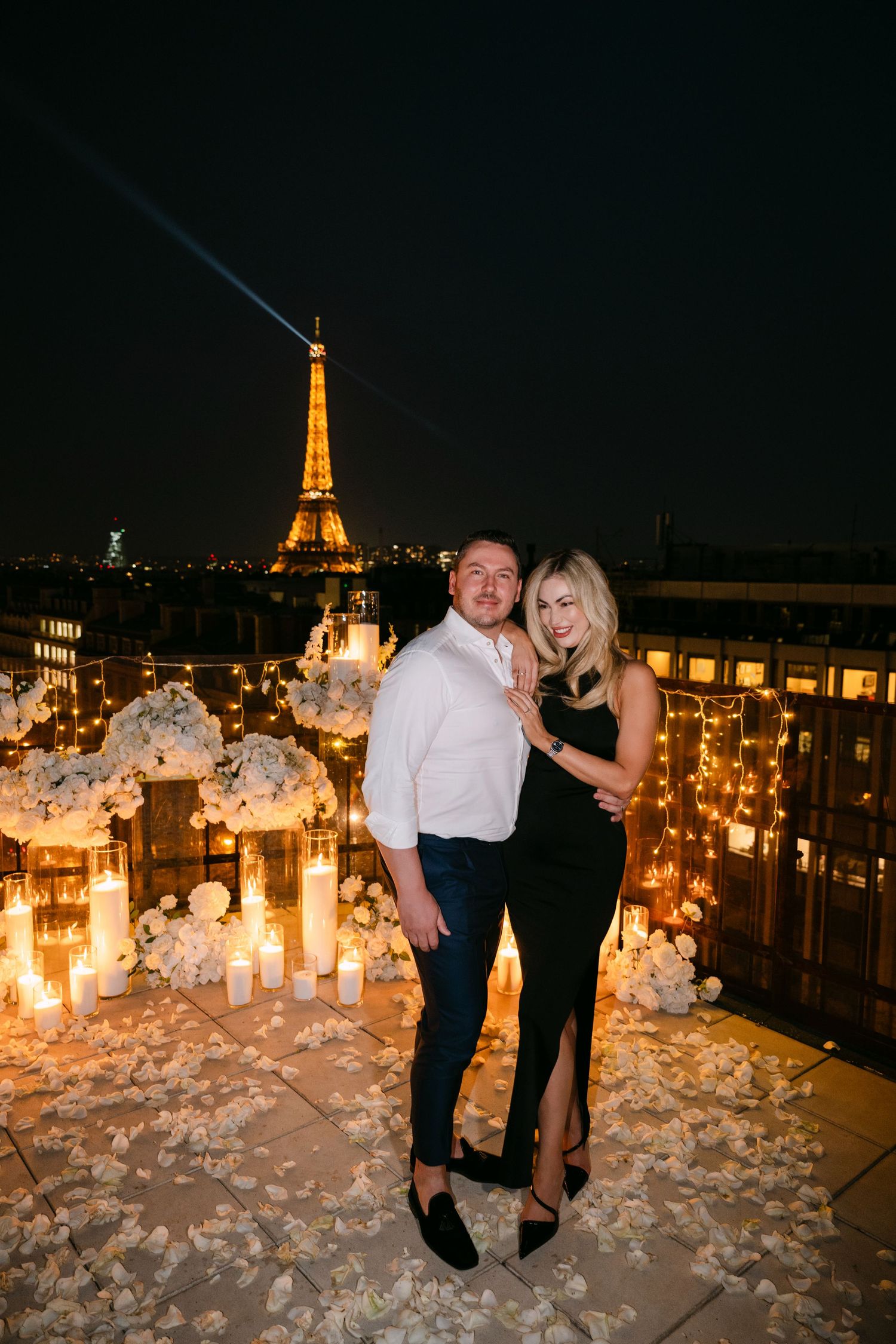Couple embraces on candlelit rooftop surrounded by white flowers with illuminated Eiffel Tower at night.
