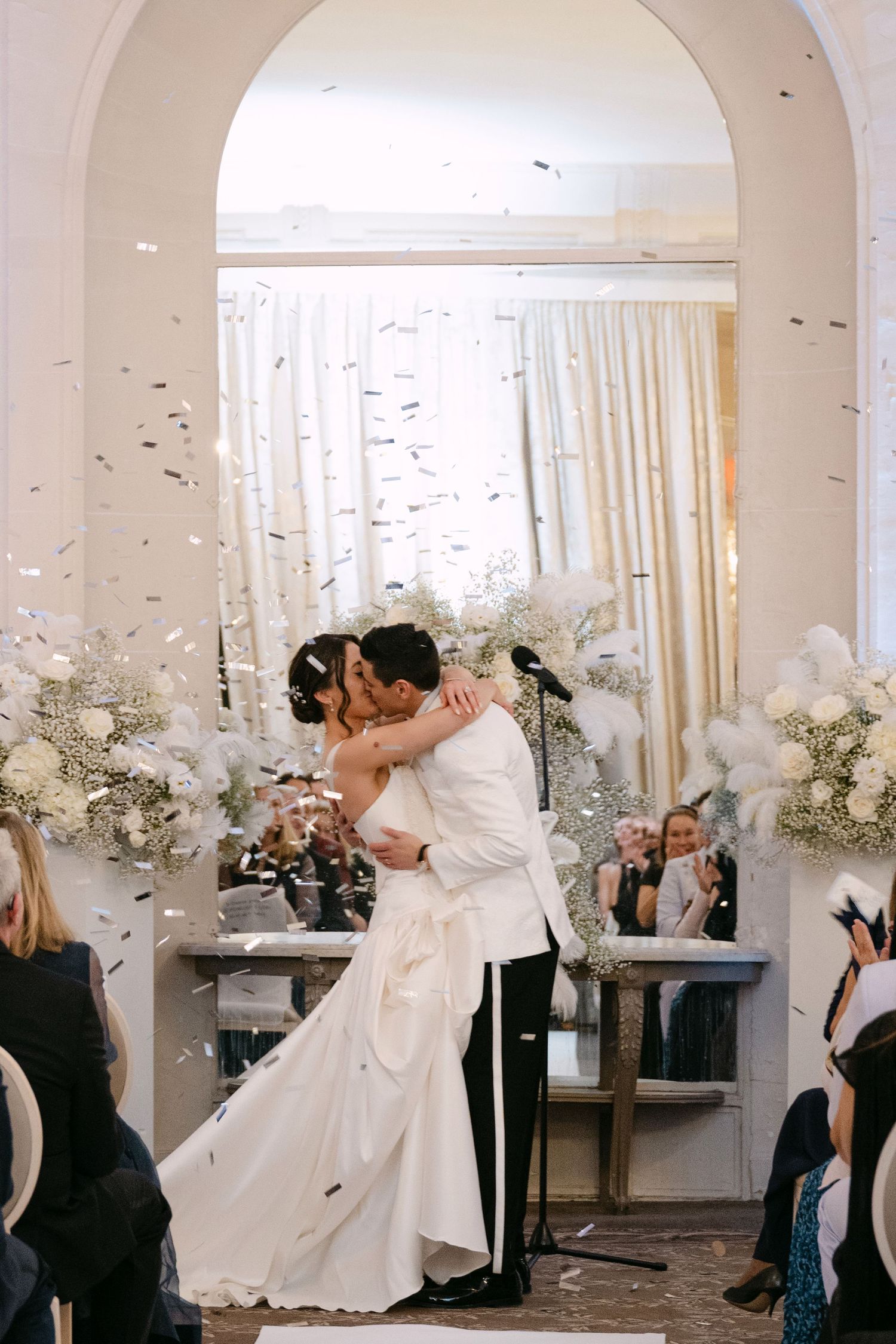 Bride and groom embrace in first kiss as confetti rains down in elegant white wedding venue.