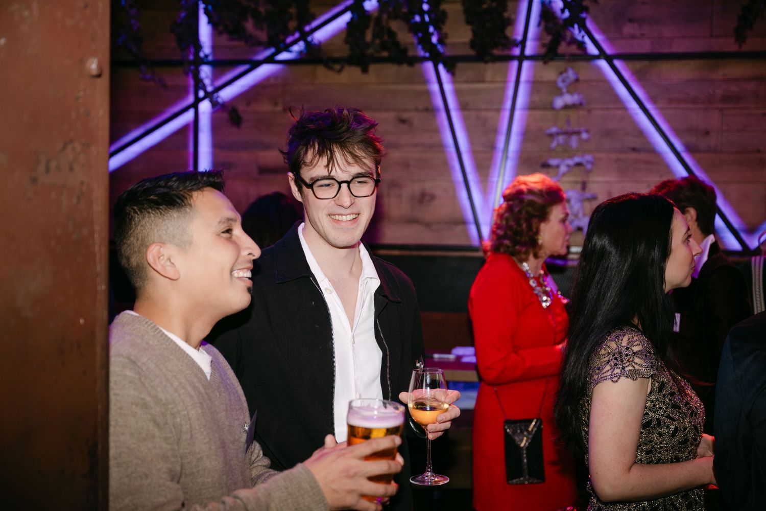 Three guests socializing and laughing at an evening party, two holding beer glasses under neon lights.