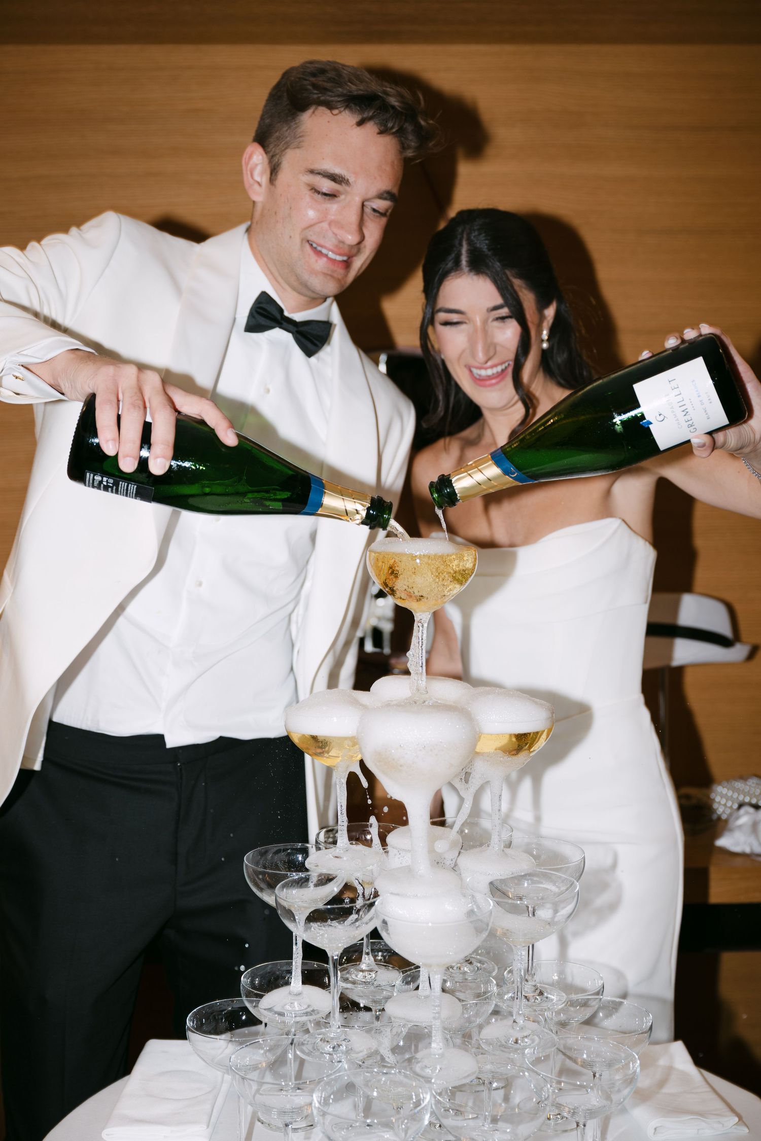 Newlyweds pouring champagne bottles over a foaming pyramid of coupe glasses at reception.