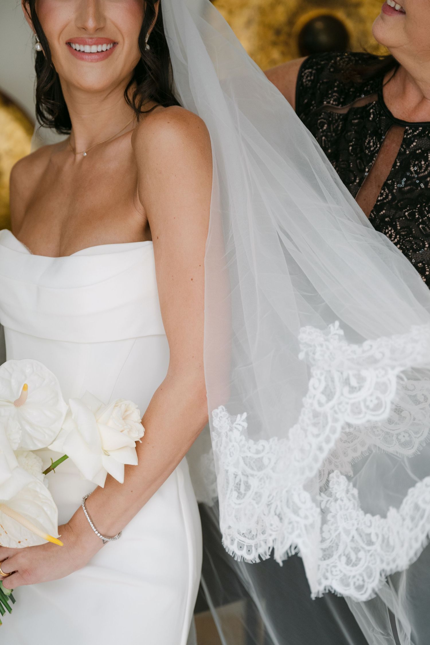 Close-up of smiling bride in strapless white gown holding white bouquet with lace veil beside her.