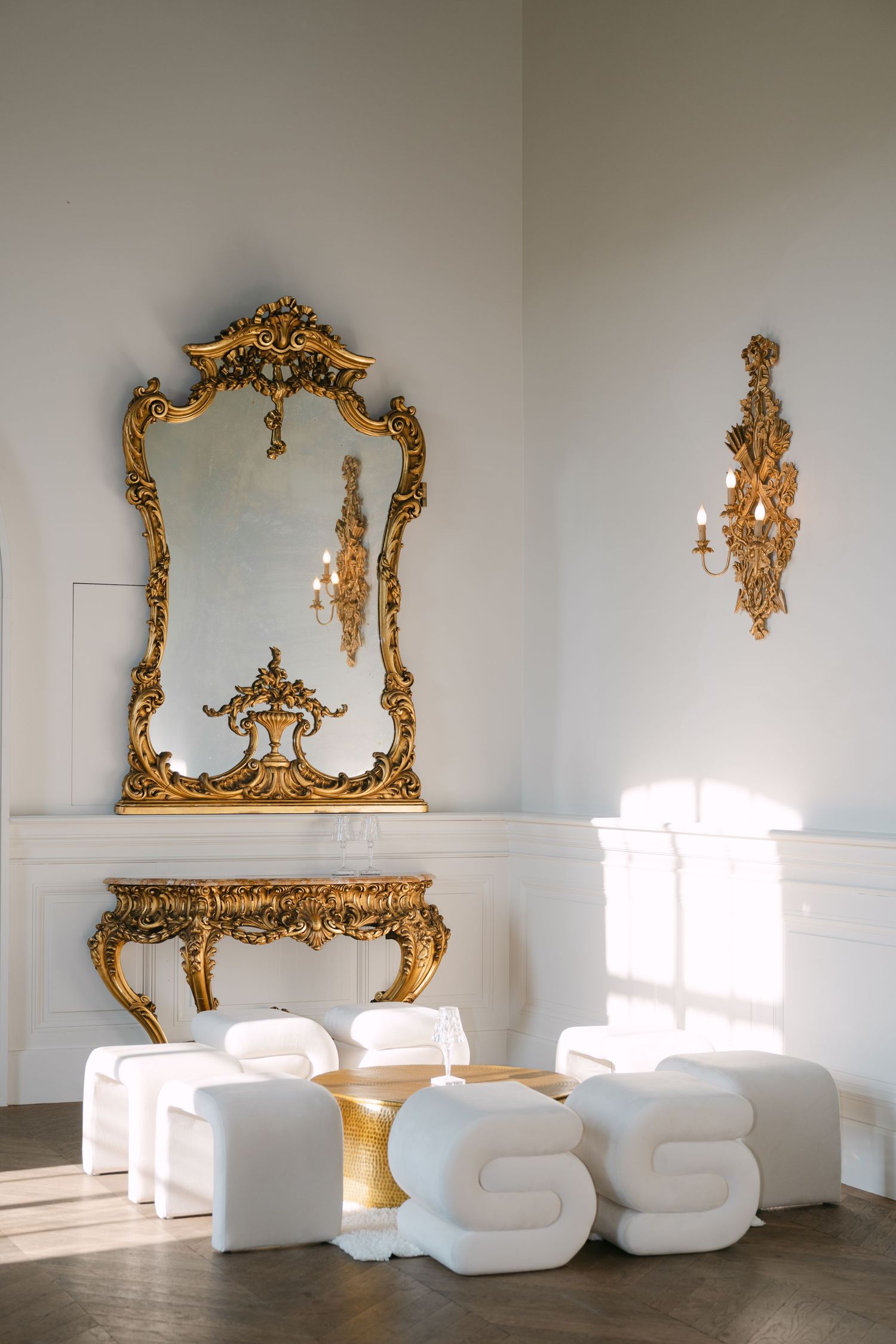Opulent room featuring a large gilded baroque mirror above a gold console table with modern white lounge chairs.