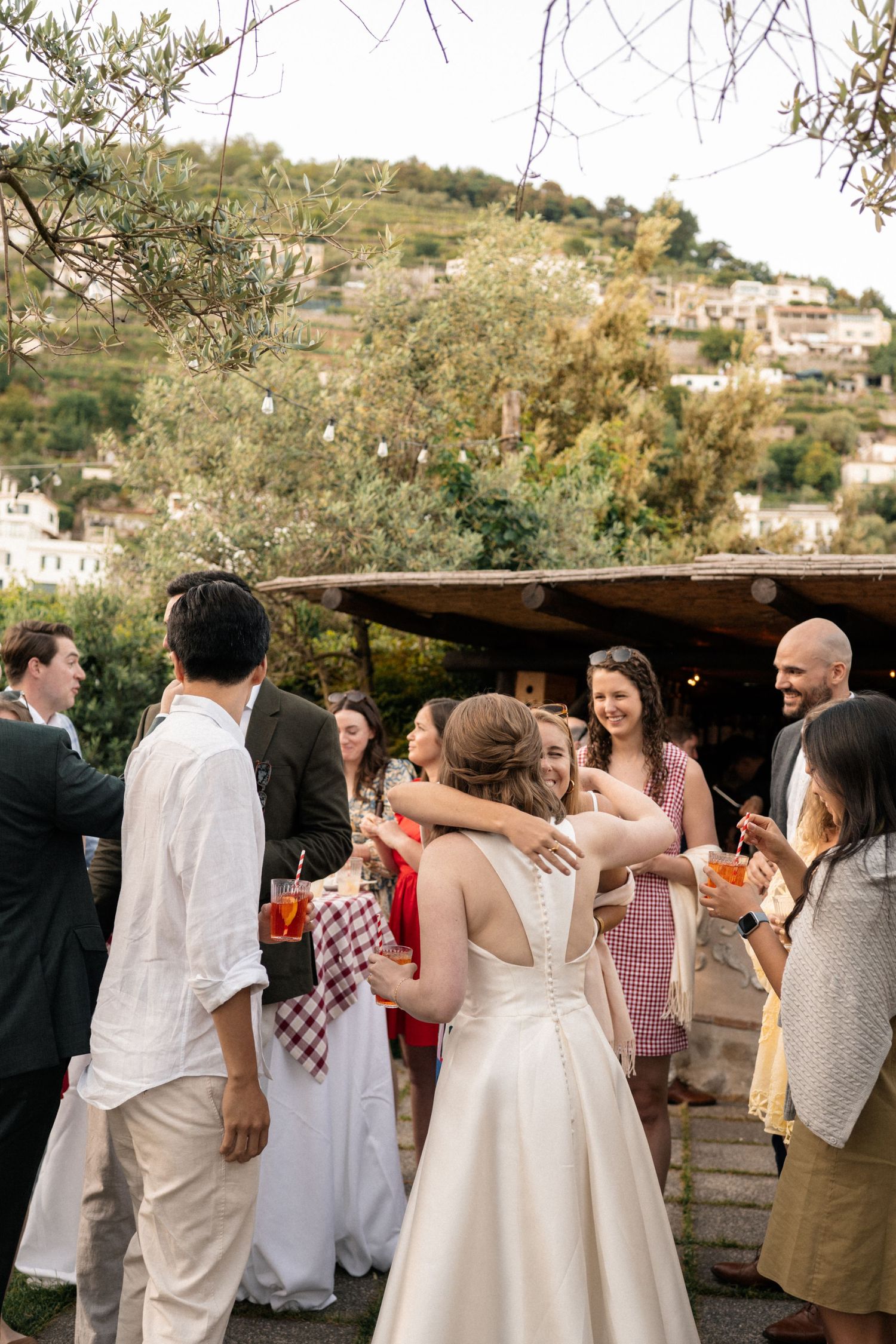 Bride in white gown greets and hugs guests at an outdoor Italian hillside wedding reception.