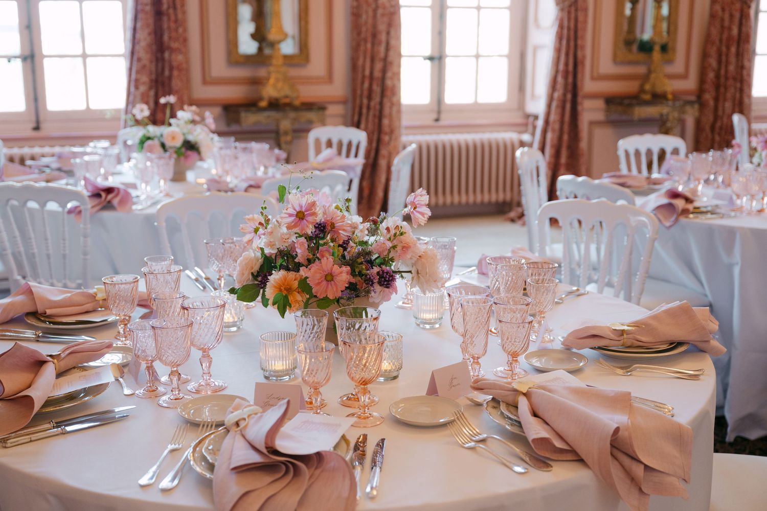 Romantic wedding reception tables with pink napkins, gold chargers, pink crystal glasses, and floral centerpieces in a chateau.