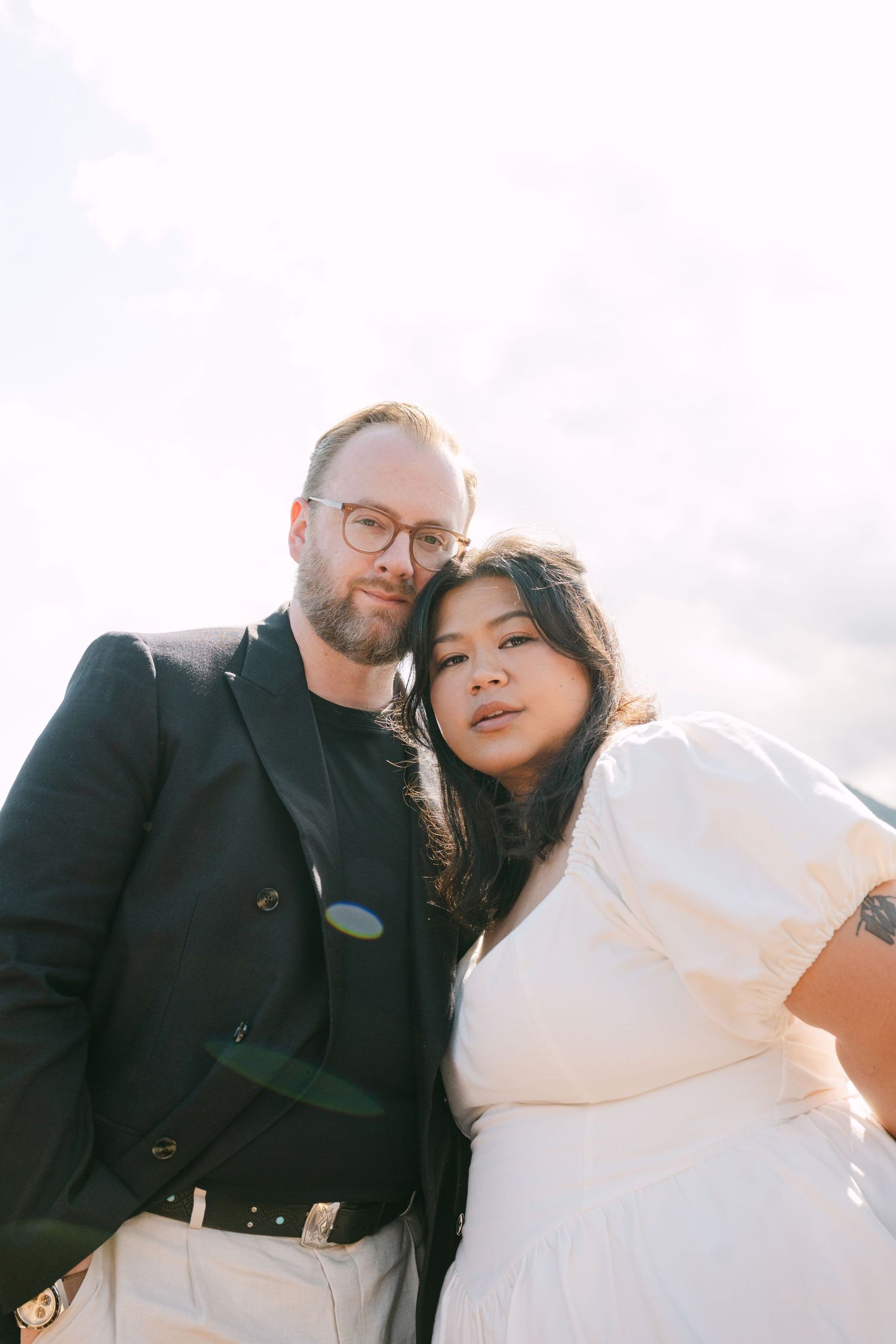 Couple in wedding attire pose together against bright cloudy sky in a low-angle intimate portrait.