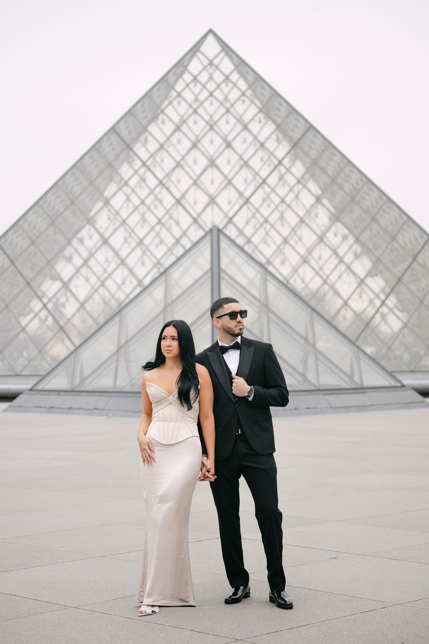 Glamorous couple posing before the Louvre glass pyramid, woman in champagne dress, man in tuxedo.