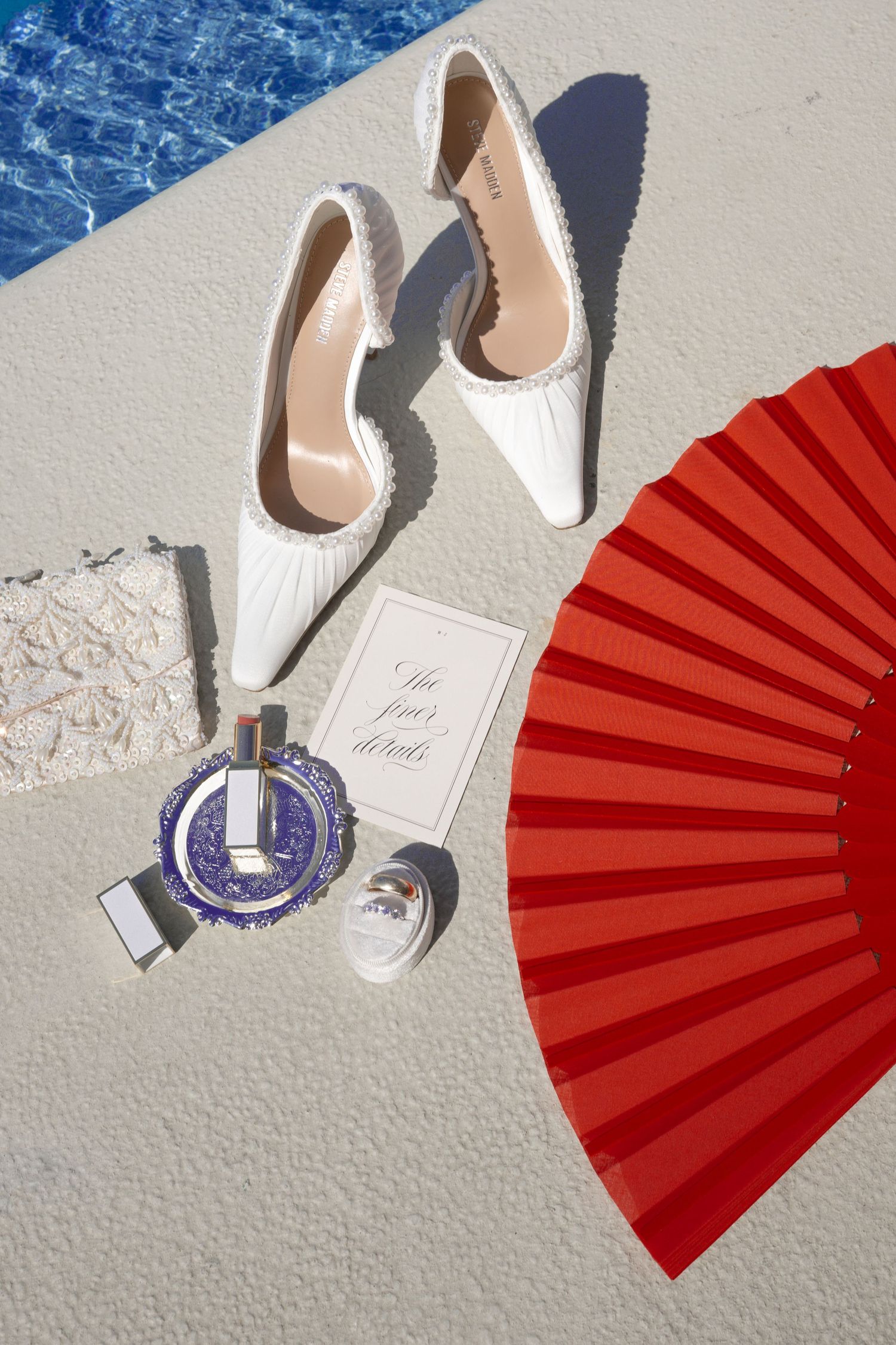 White bridal heels, red folding fan, perfume bottles, and wedding details card arranged near a pool.