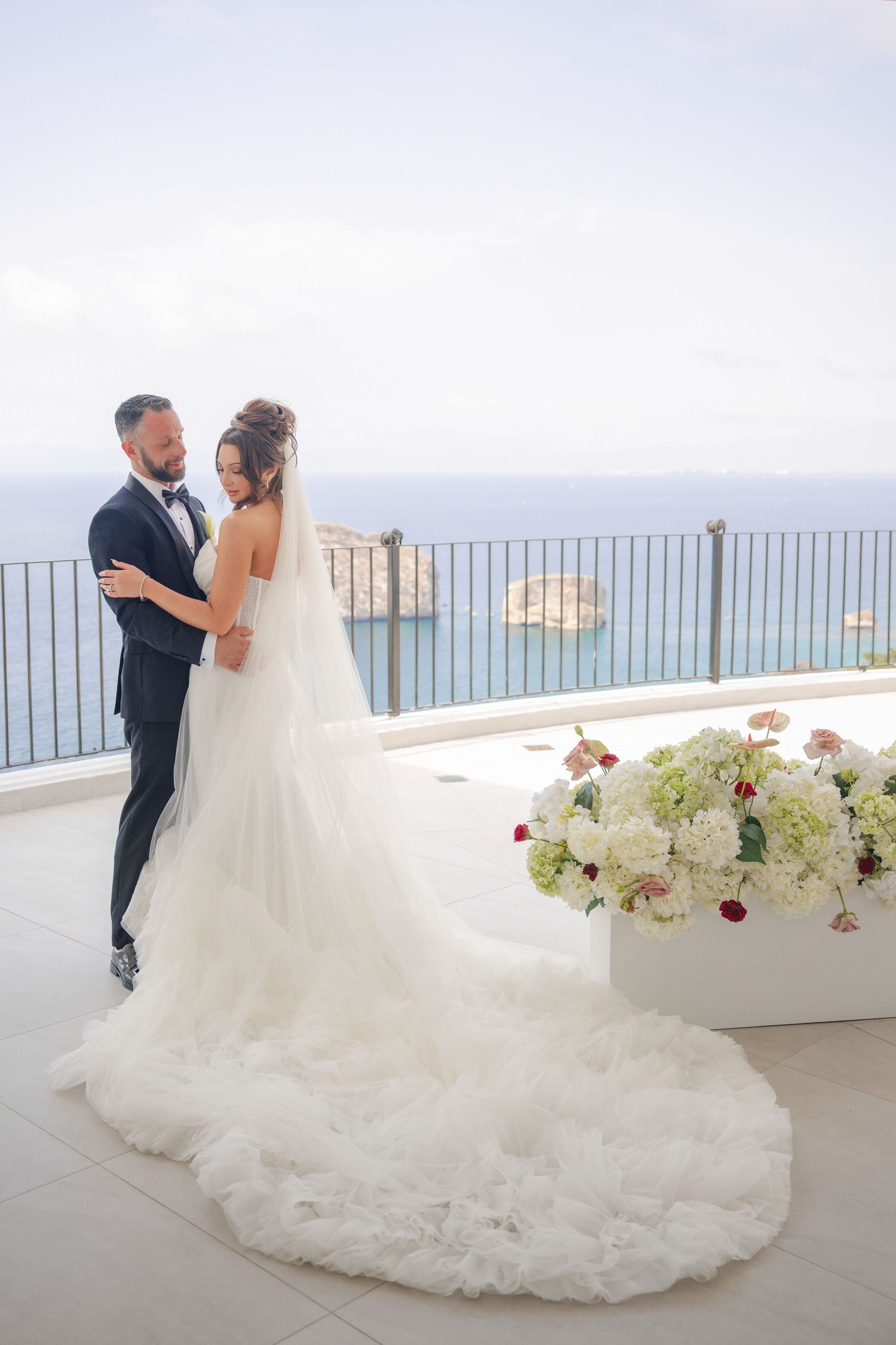 Bride and groom embrace on a seaside terrace with ocean views, white floral arrangements, and a flowing cathedral veil.