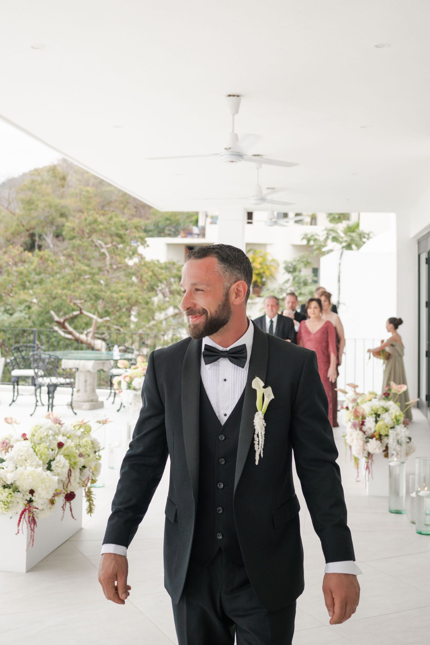 Smiling groom in black tuxedo with bow tie walks down aisle with wedding party following behind.