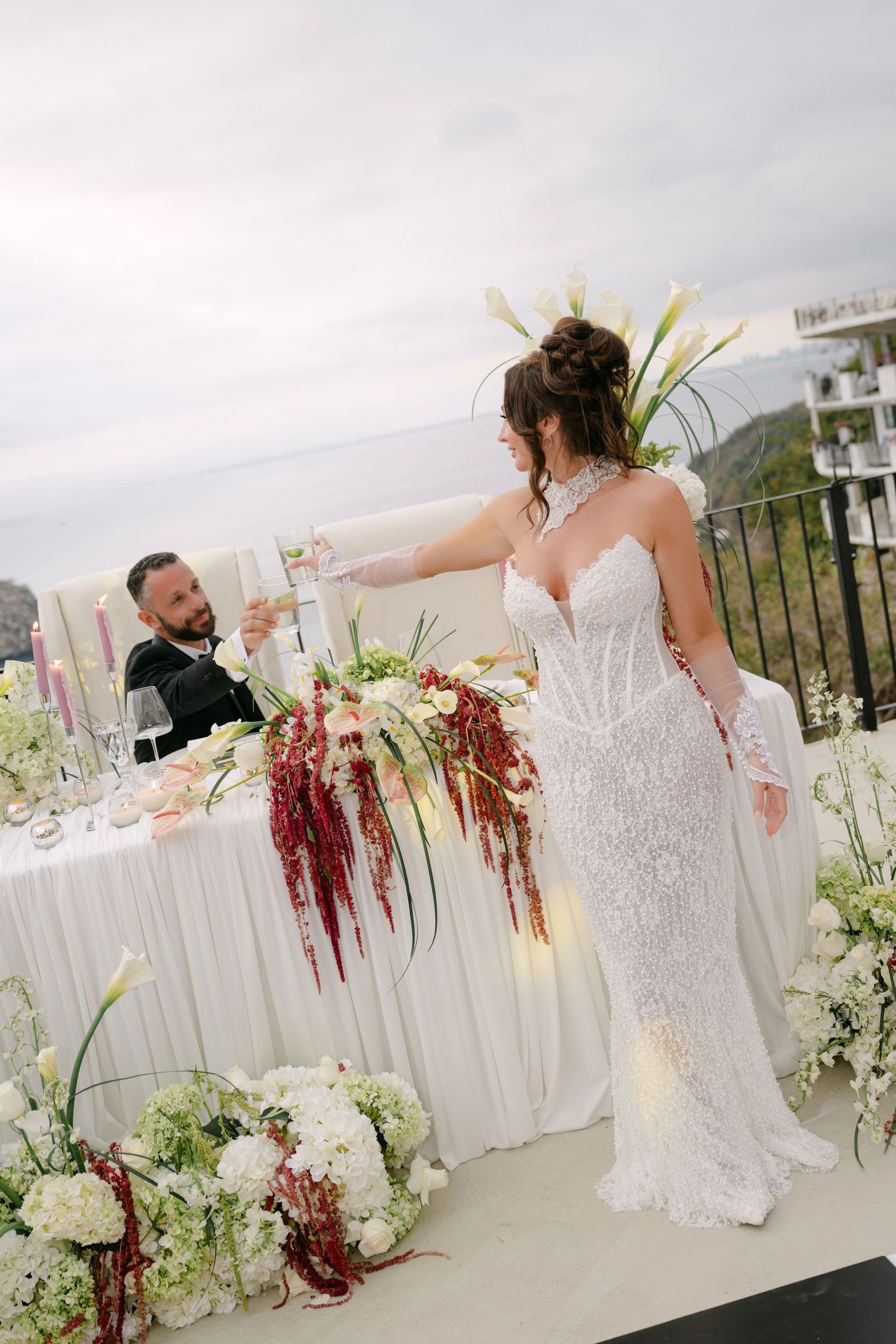 Bride in lace gown toasting with groom at elegant outdoor wedding reception with floral arrangements.