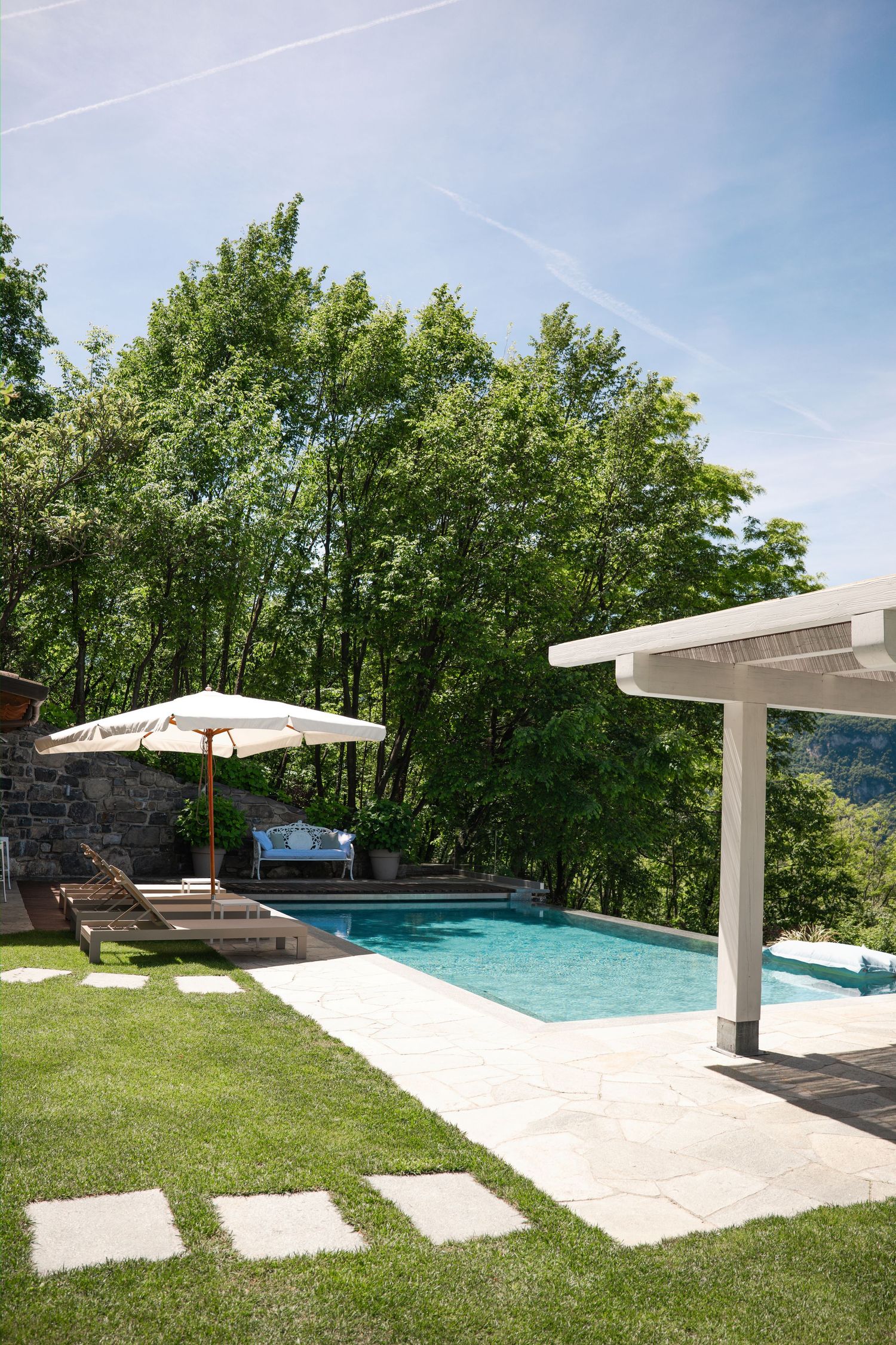 Luxury outdoor pool with lounge chairs, white umbrella, pergola, and lush green trees under a clear blue sky.