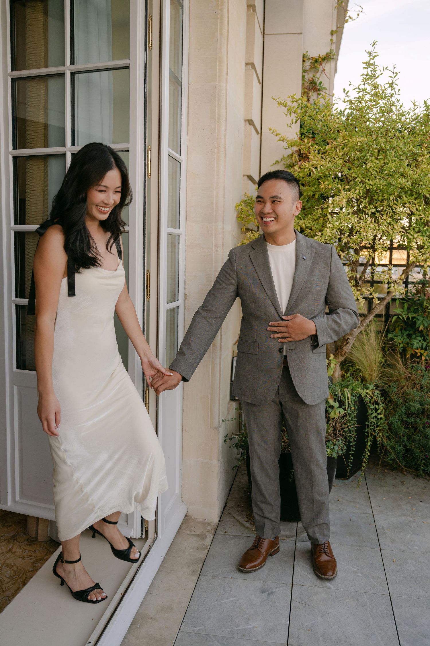 Smiling couple holding hands on a sunlit terrace, she in a white satin slip dress and he in a gray suit, beside open French doors.