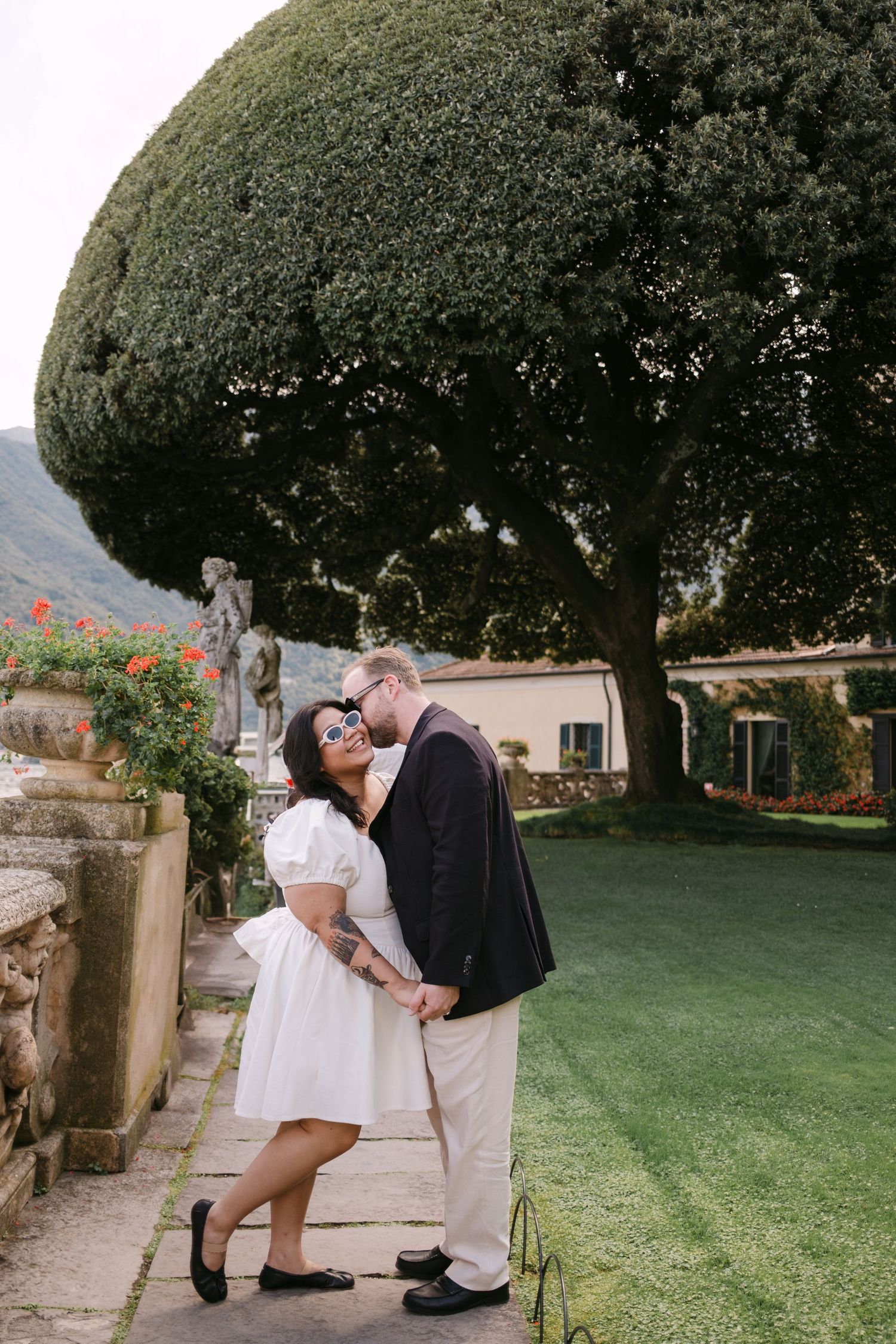 Couple sharing a romantic kiss in an Italian garden with sculpted trees and stone statues.