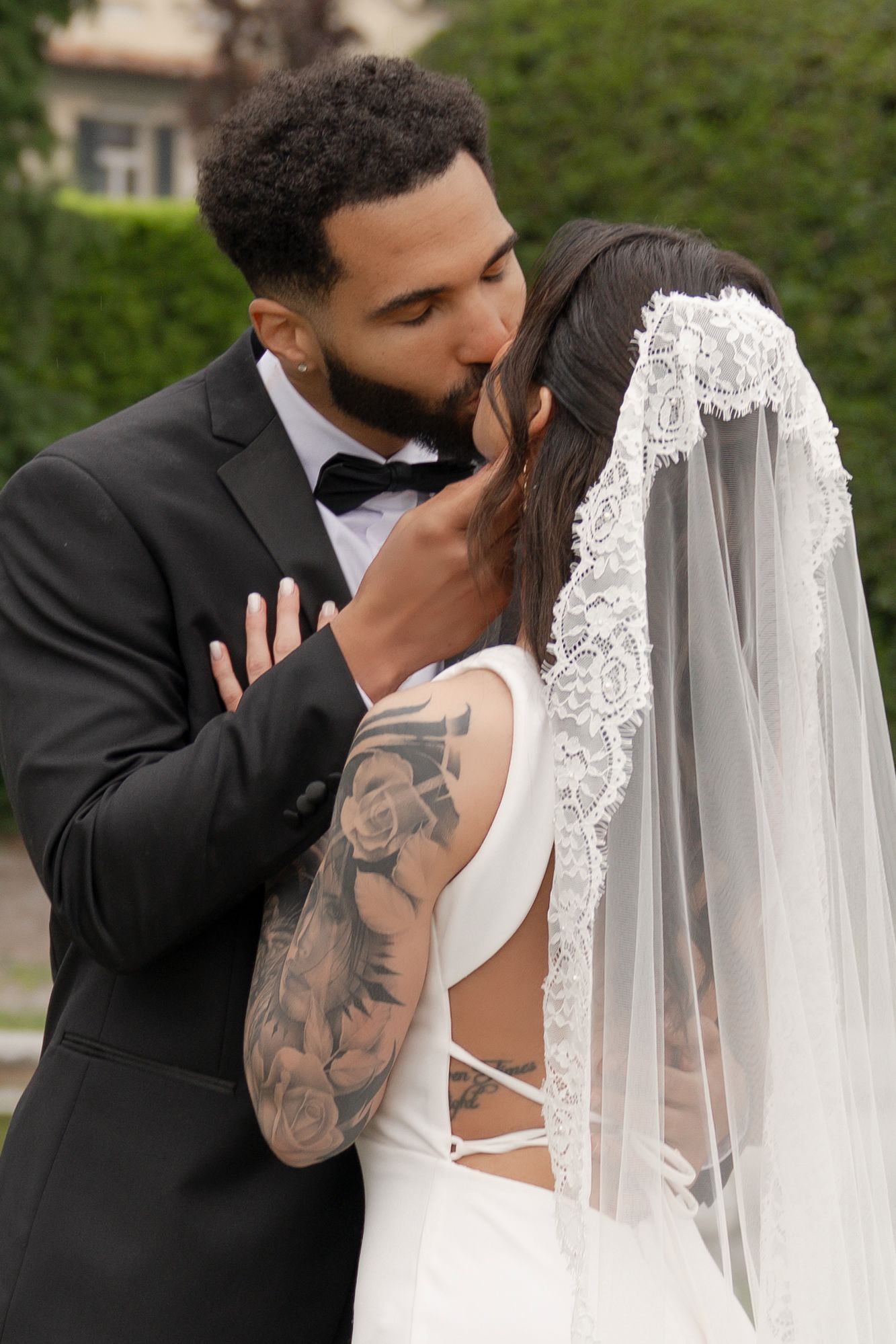 Groom in black tuxedo kissing tattooed bride in white dress and lace veil outdoors at wedding ceremony.
