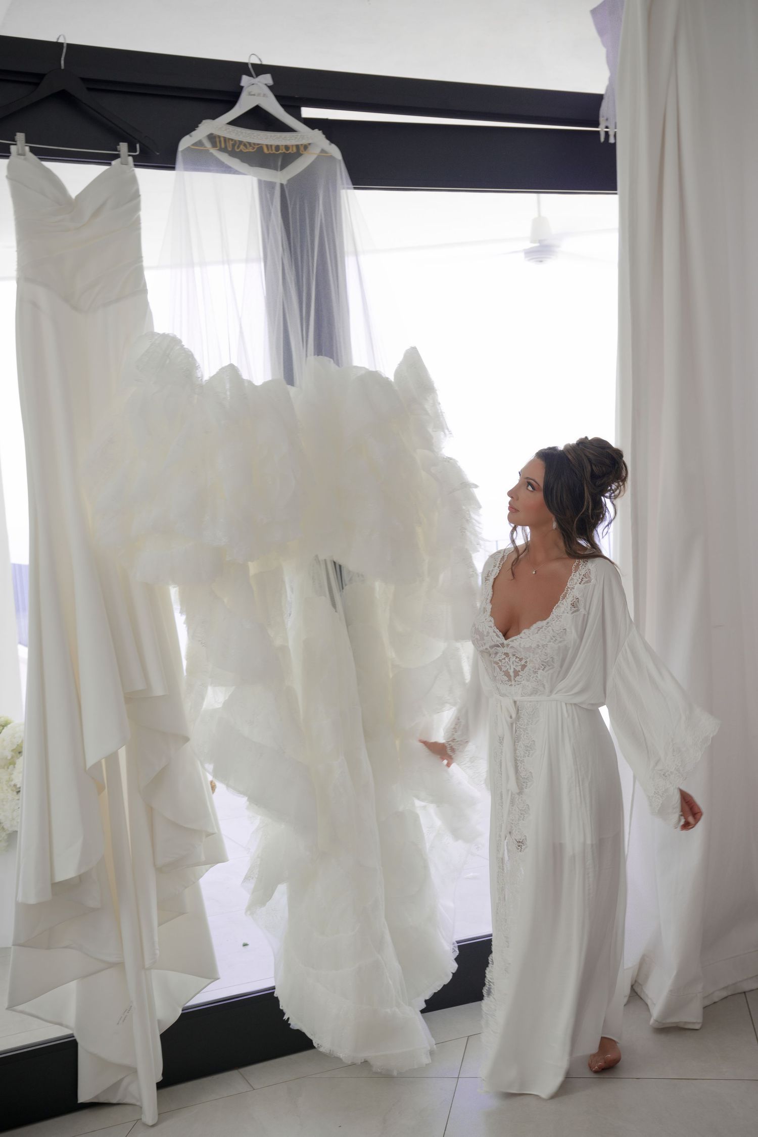 Bride in white robe admiring her elegant ruffled wedding gown hanging near a bright window.