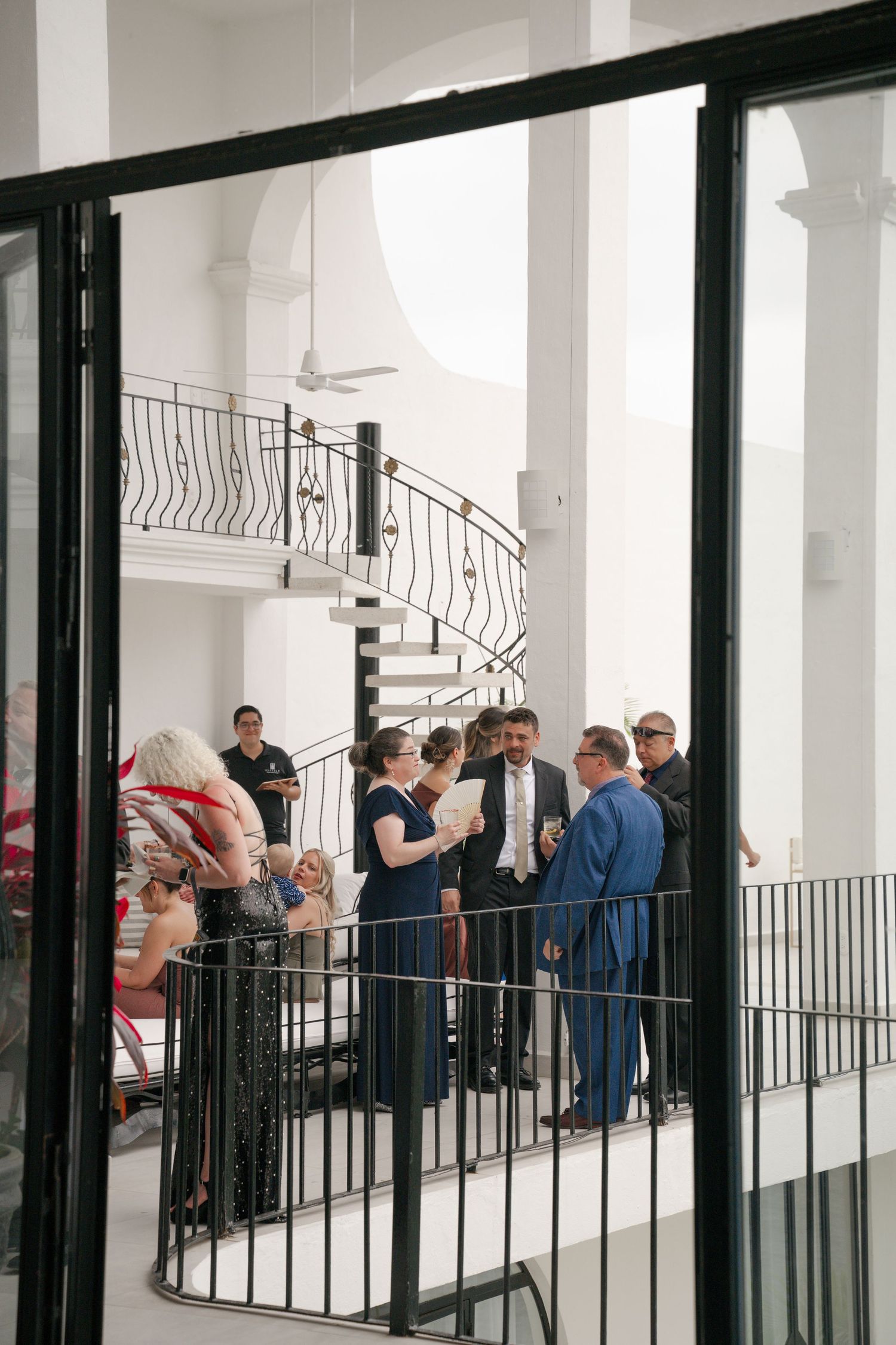 Elegantly dressed wedding guests mingle on a balcony inside a white venue with a spiral staircase.