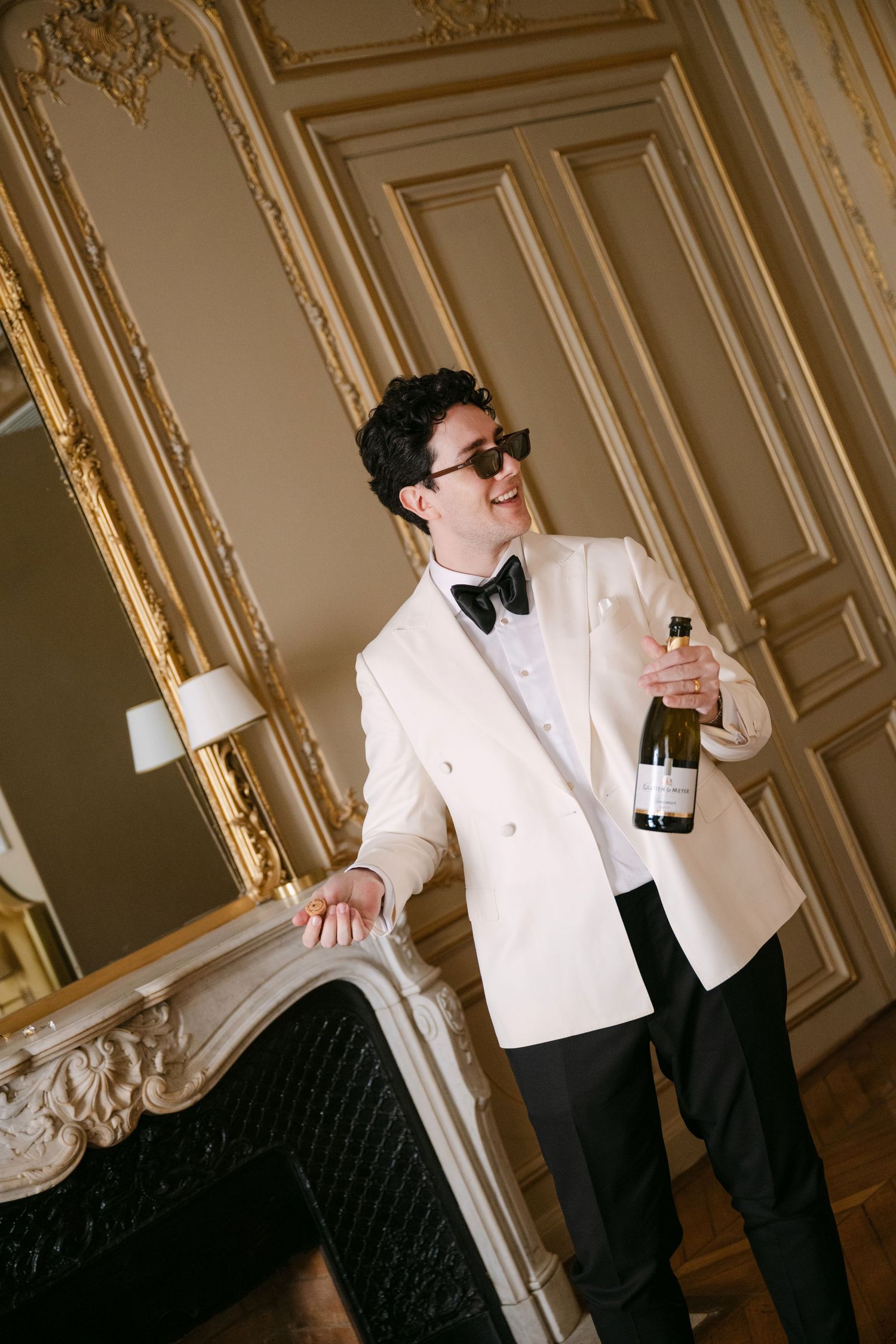 Groom in white tuxedo jacket and black bow tie holds champagne bottle by an ornate fireplace.