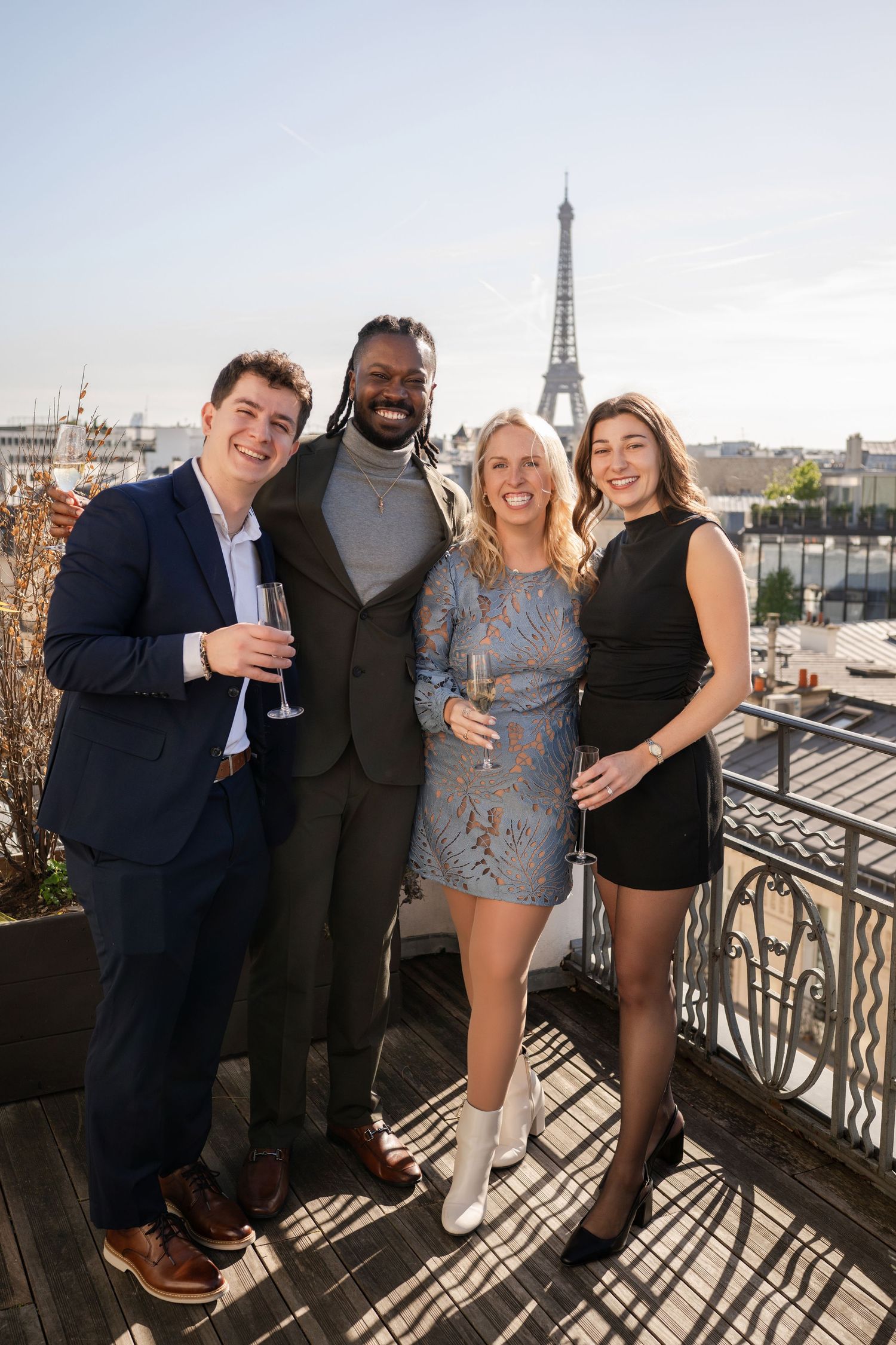 Four friends celebrating with champagne on Paris rooftop terrace with Eiffel Tower visible behind them.