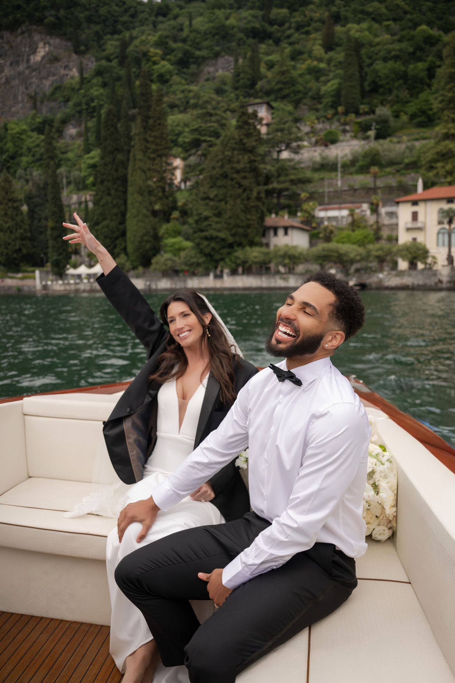 Joyful groom laughing on luxury boat with bride wearing wedding dress and black blazer on lake.