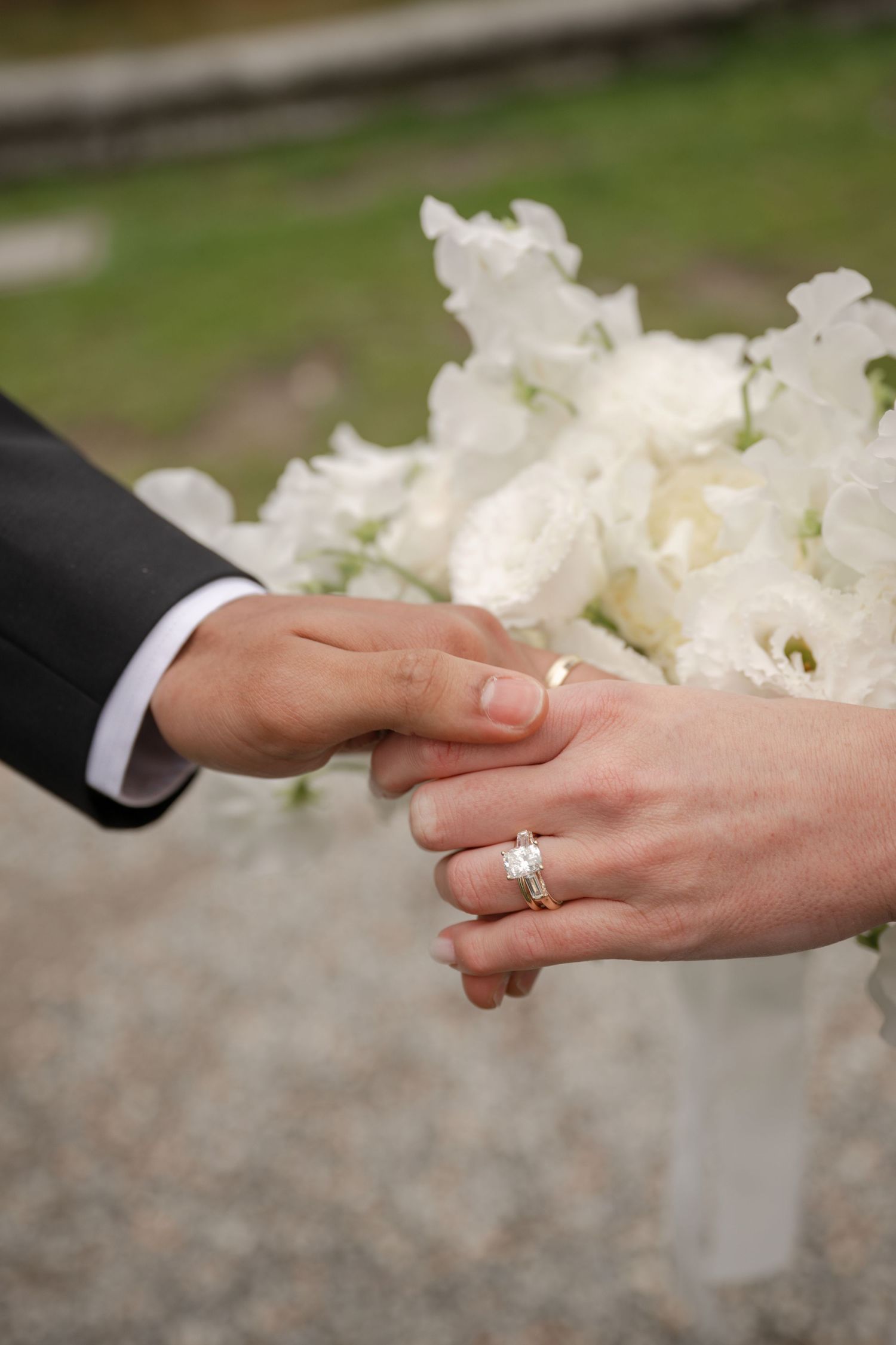 Bride and groom holding hands showing diamond engagement ring with white floral bouquet at wedding ceremony.