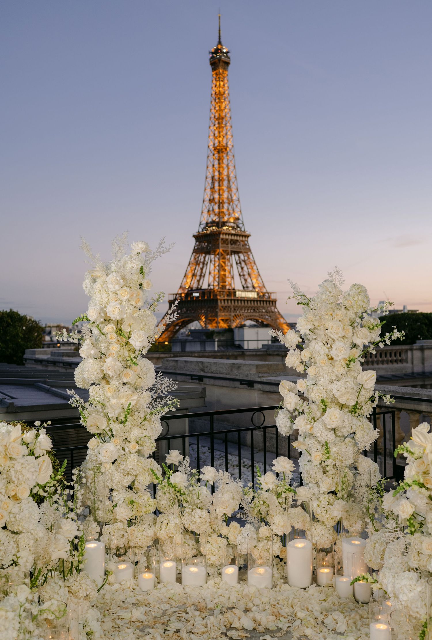 Eiffel Tower glows at dusk behind towering white floral columns and candles at an elegant Paris rooftop proposal.