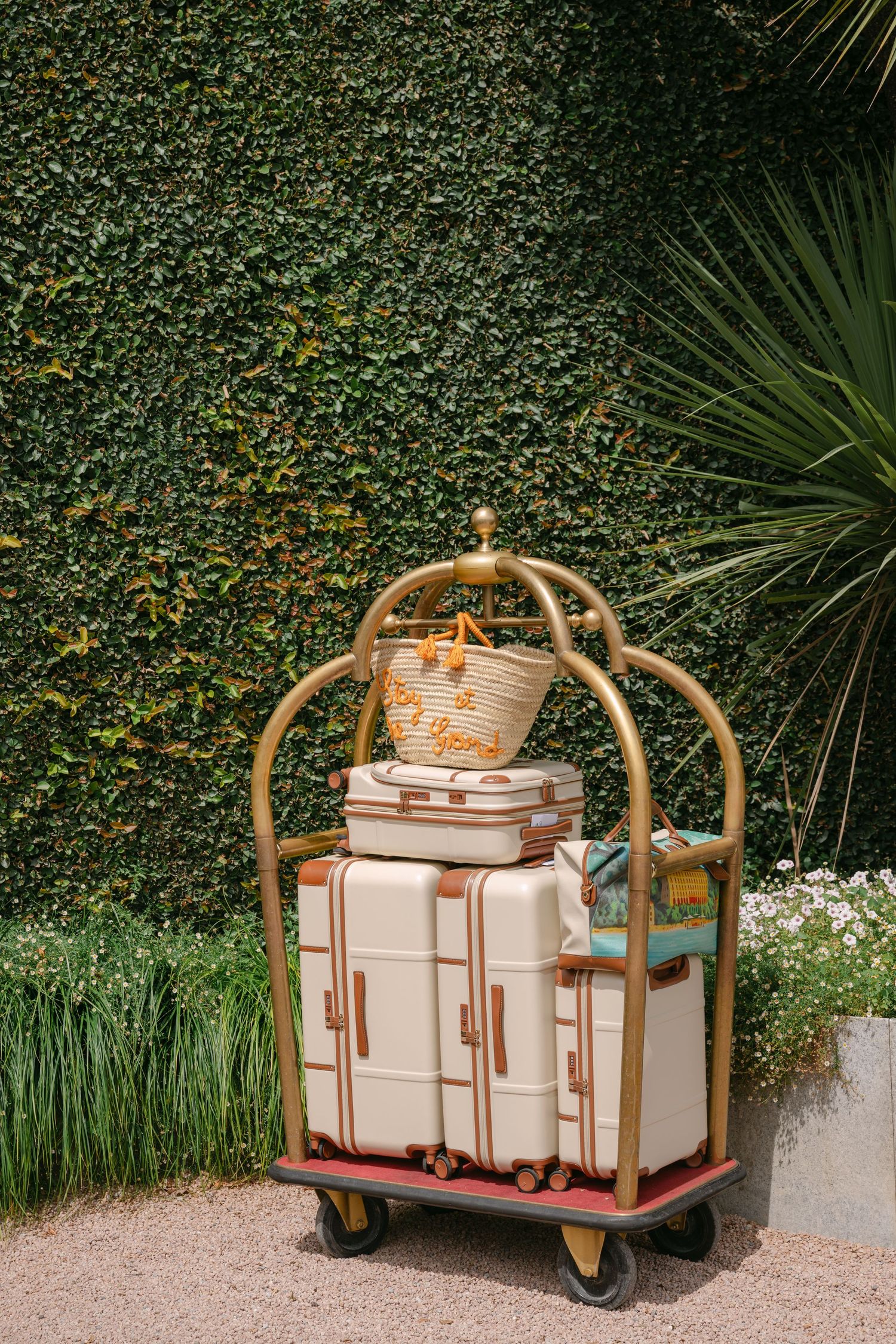 Gold hotel luggage cart with cream suitcases and wicker basket against lush ivy wall.