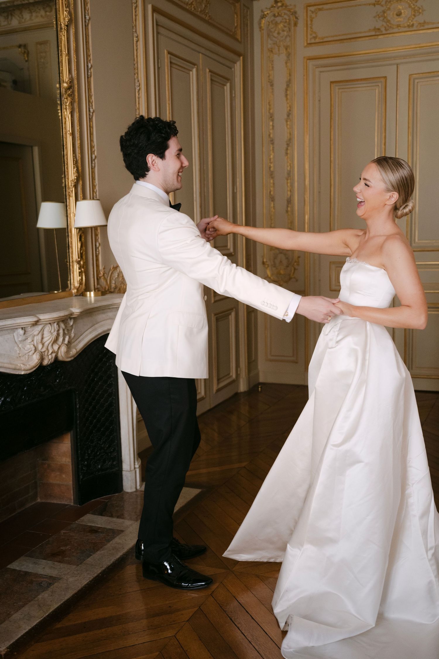 A couple in wedding attire dances and twirls together in an elegant room with ornate gold-trimmed walls.
