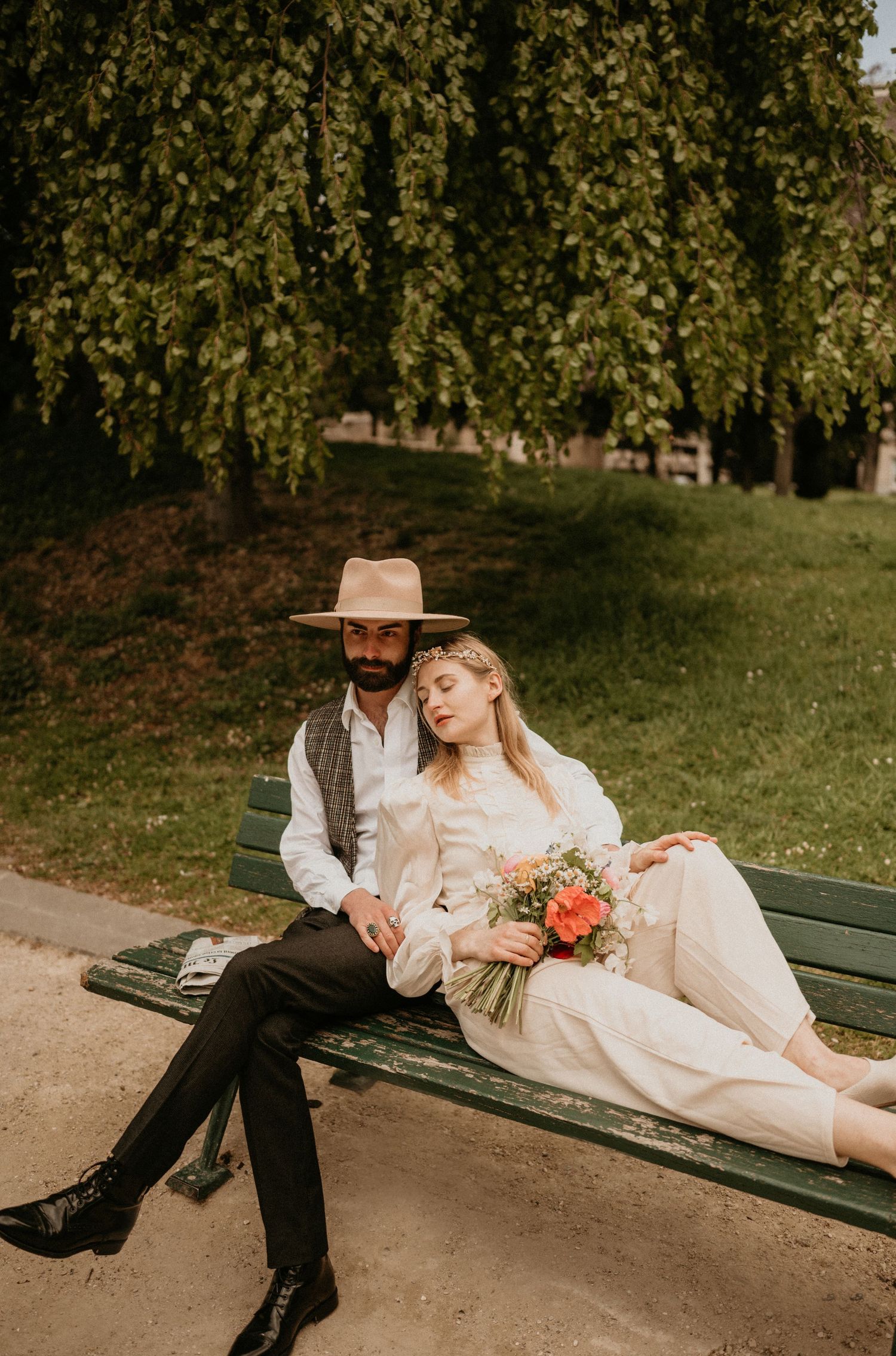 Couple lounging on green park bench with floral bouquet under weeping willow.