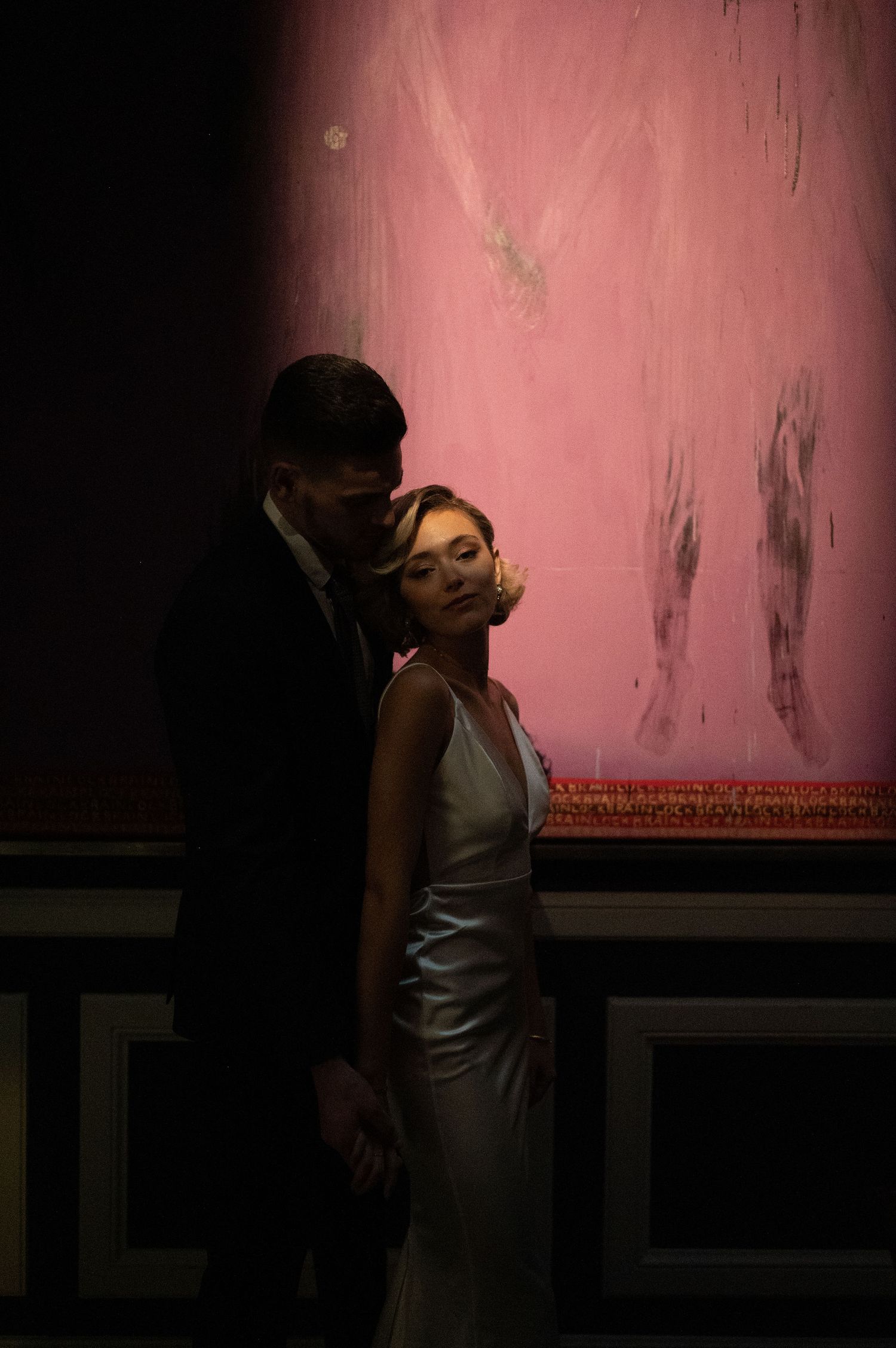 Romantic couple pose against pink wall in formal attire with dramatic lighting.