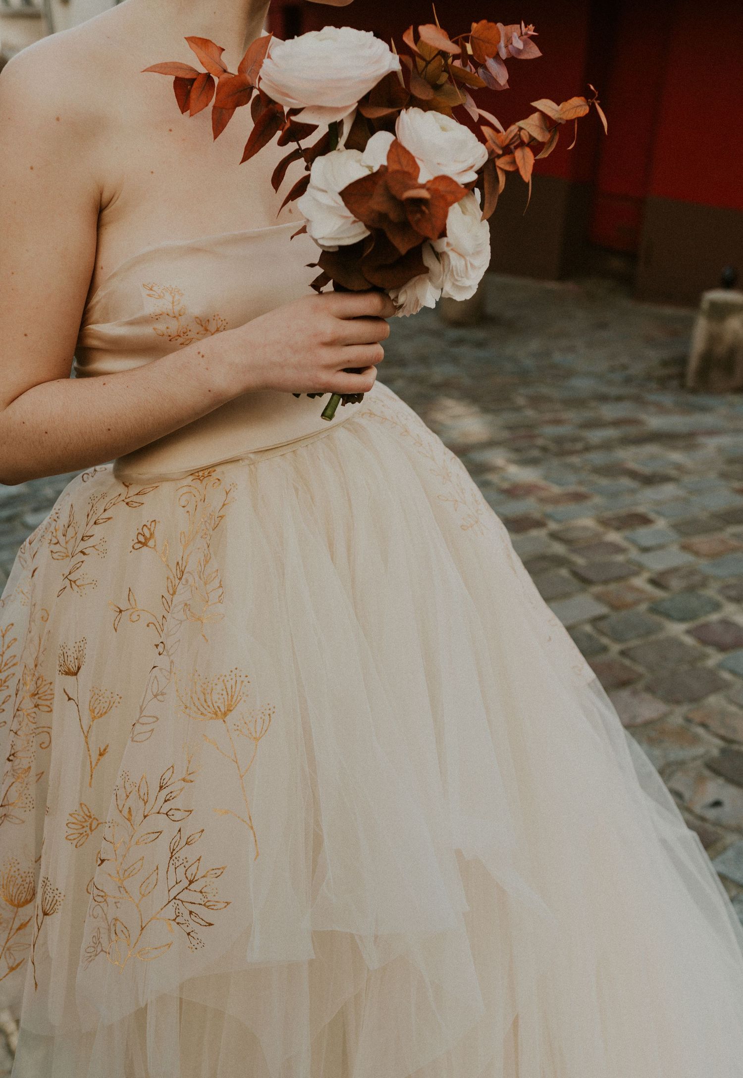 Close-up detail of bride holding autumn bouquet against champagne colored wedding dress with floral details.