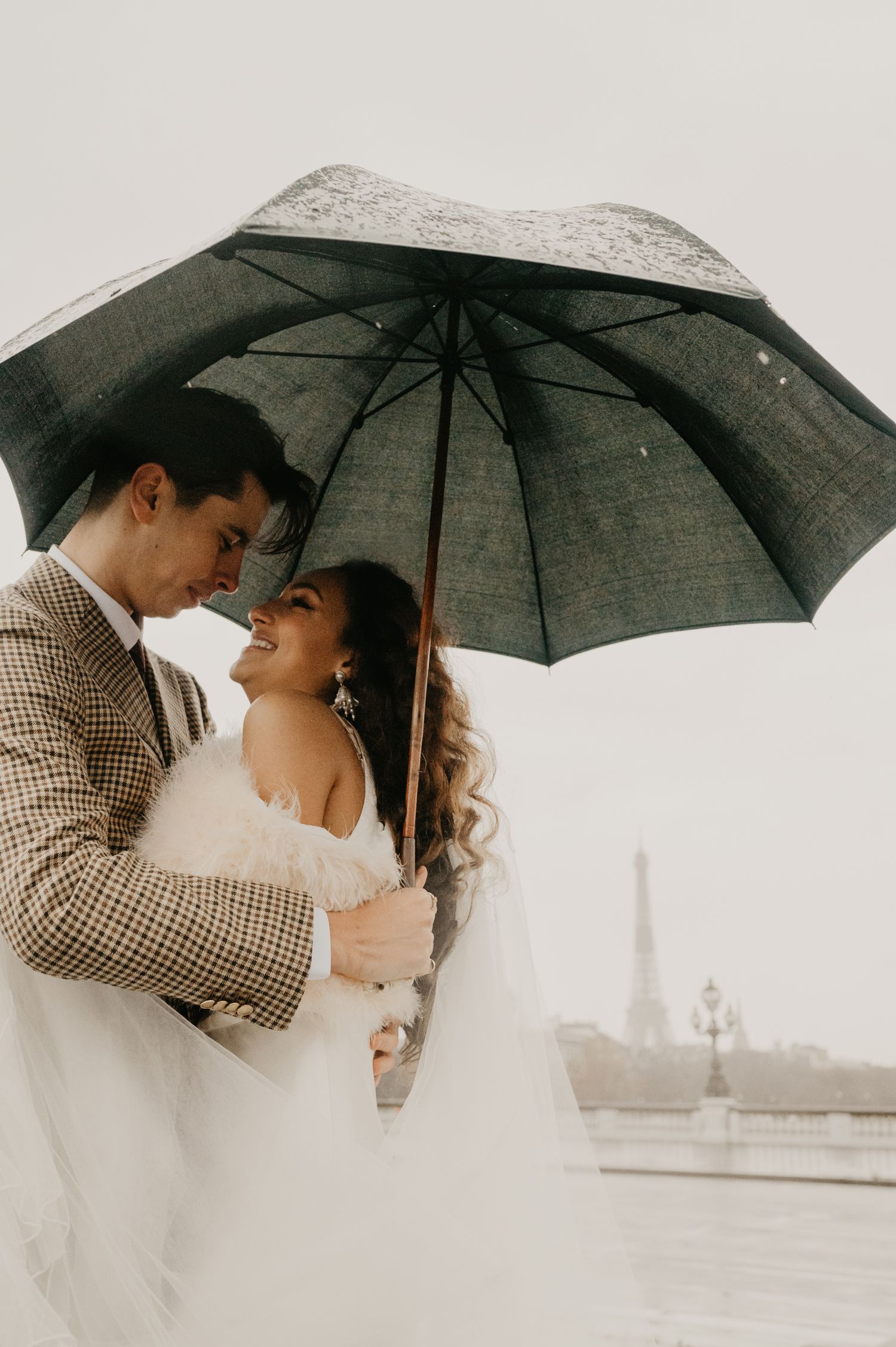 Romantic couple under black umbrella with Eiffel Tower visible in soft sepia tones.