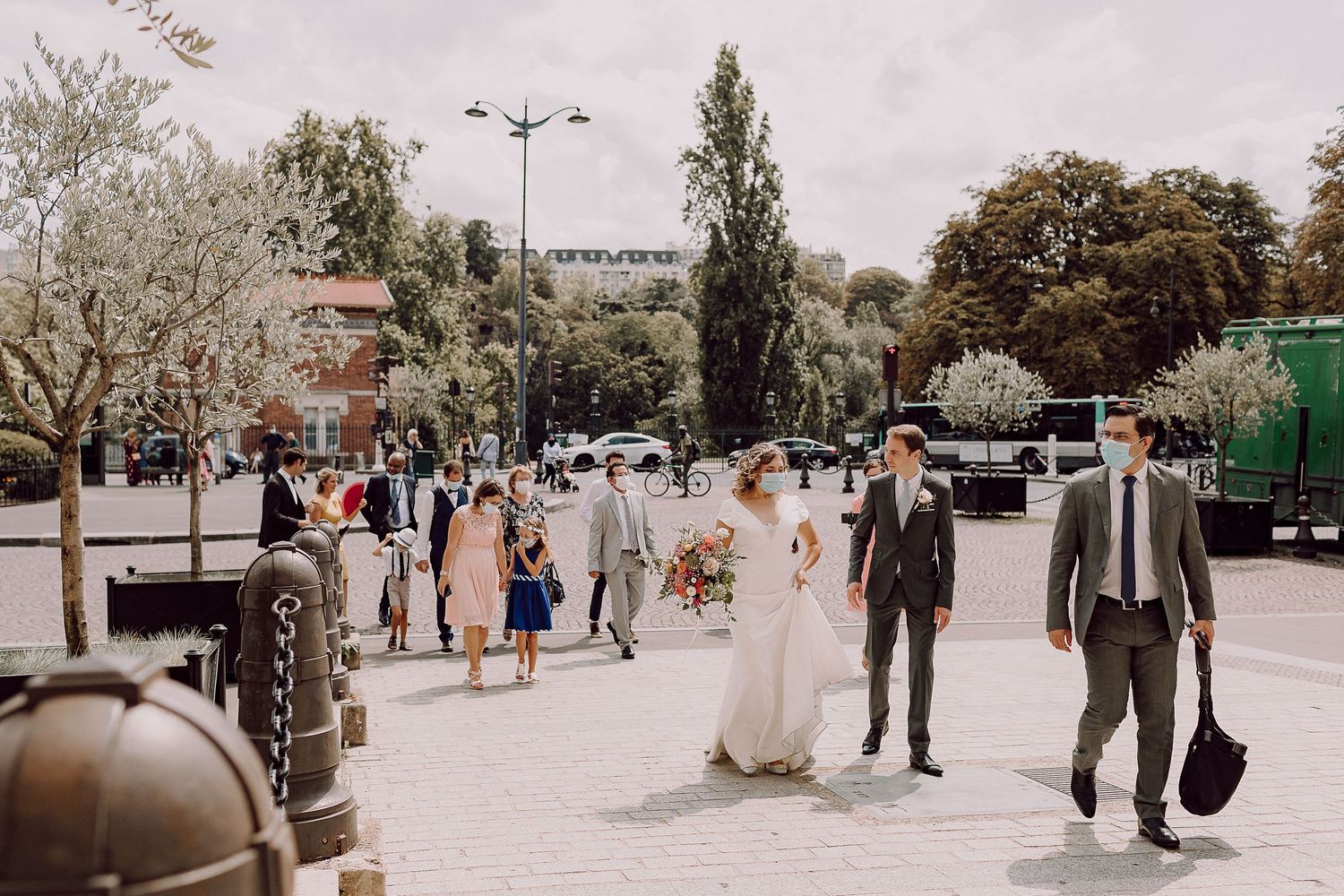 Wedding party walking together on a street with sunlit trees and parked cars in the background.