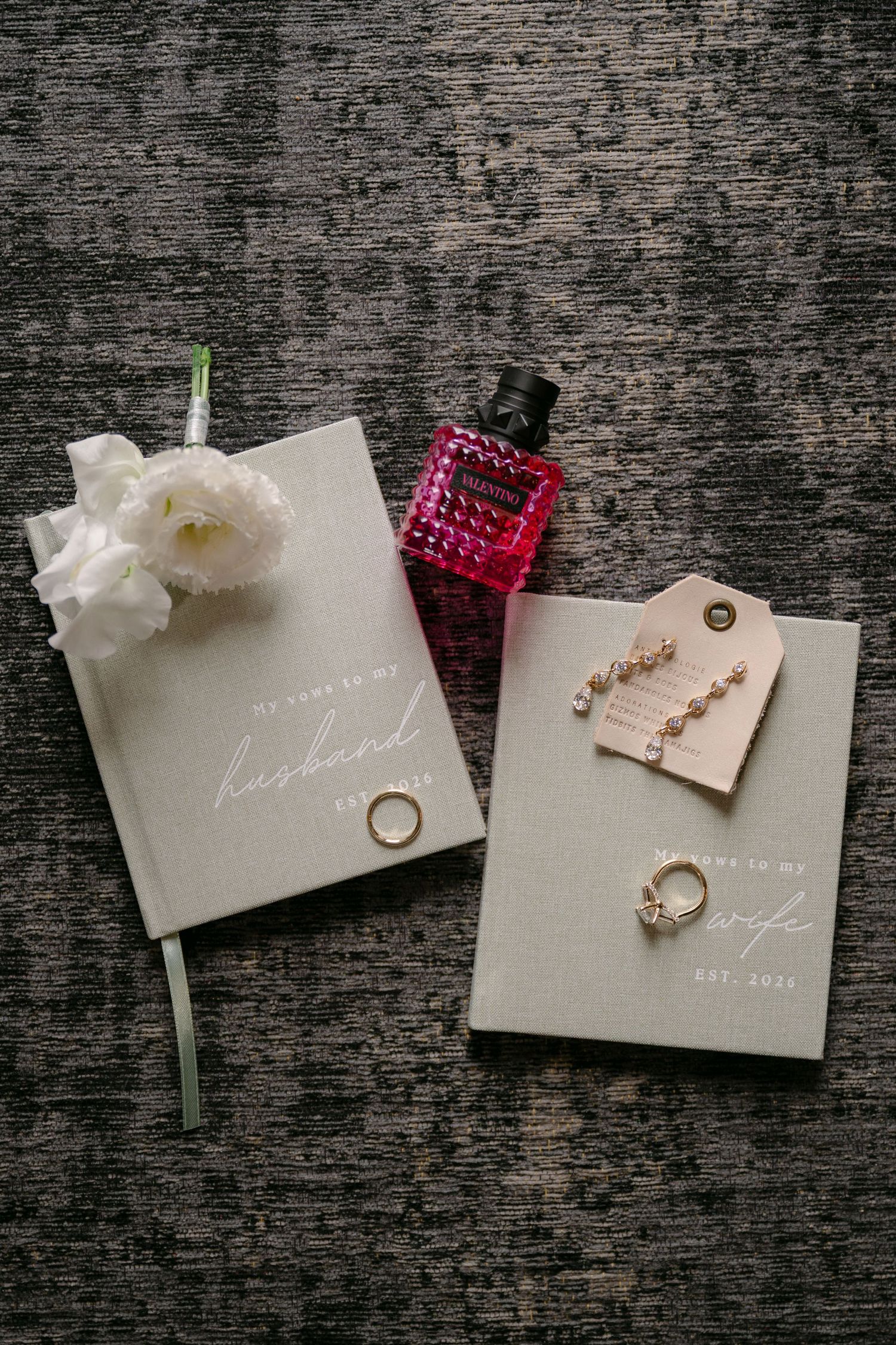 Wedding flat lay with vow books, diamond rings, drop earrings, white flowers, and red perfume bottle on gray texture.