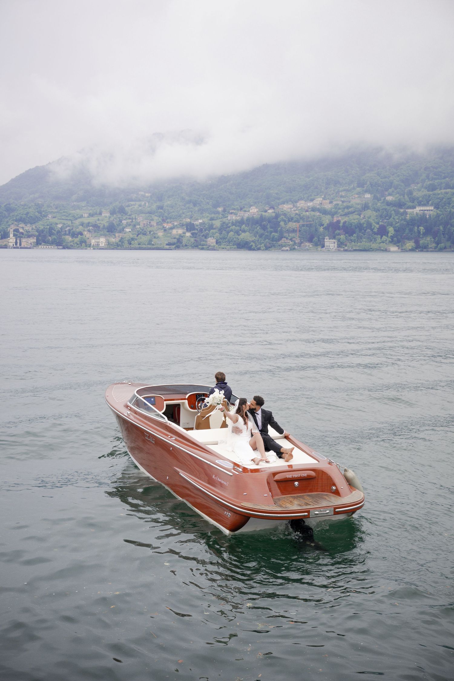 Bride and groom riding a classic wooden speedboat on a misty lake with green hills in the background.