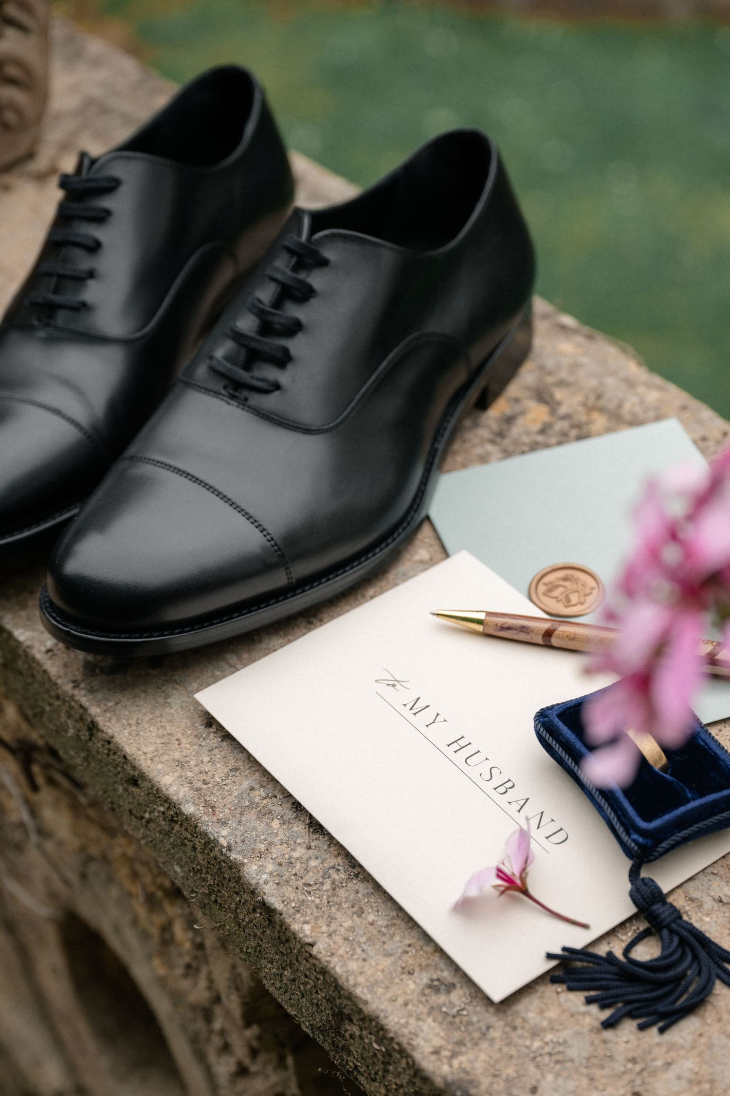 Wedding flat lay with black Oxford shoes, To My Husband vow book, gold pen, wax seal, and ring box.