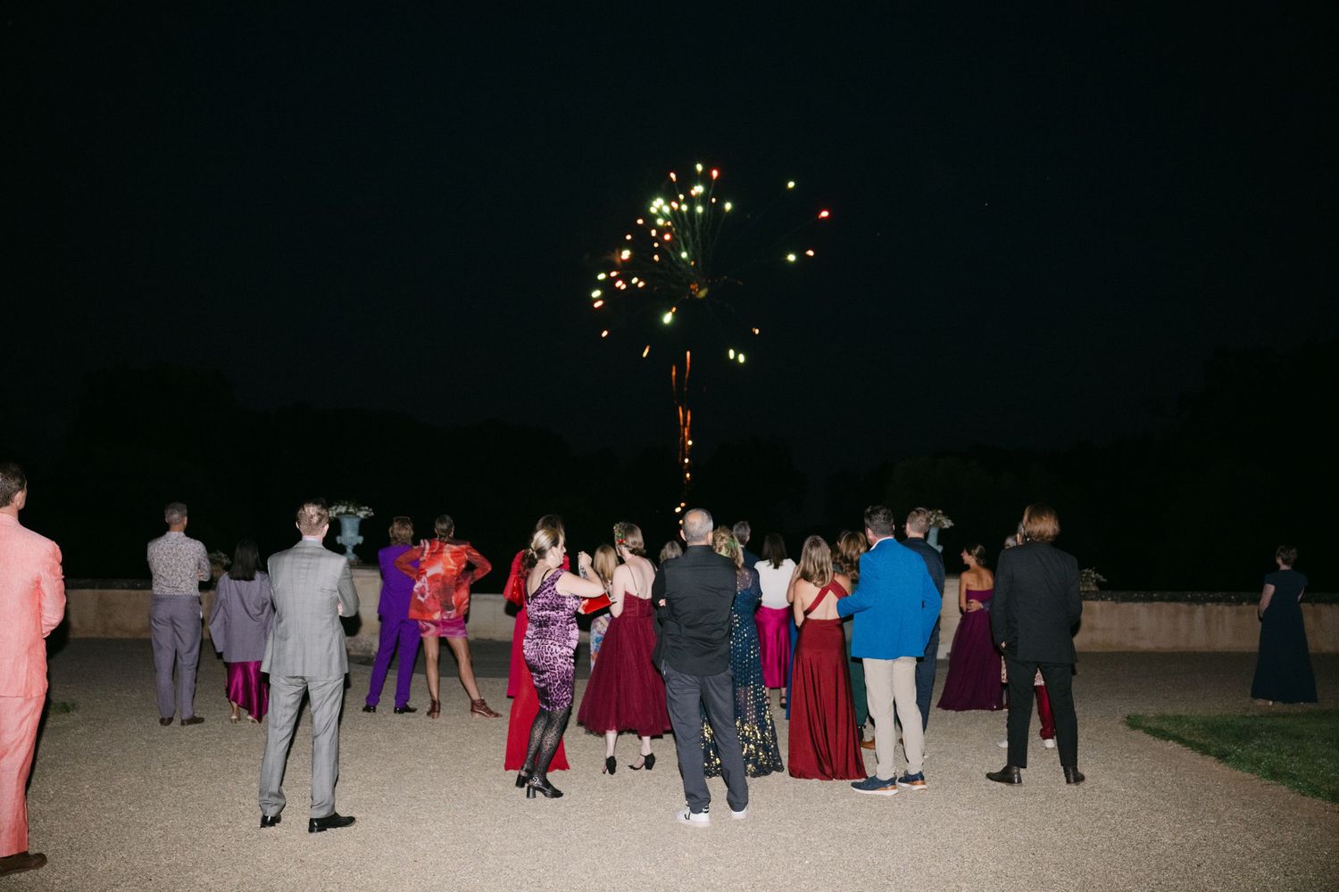 Elegantly dressed wedding guests watching a sparkling firework display on an outdoor gravel terrace.
