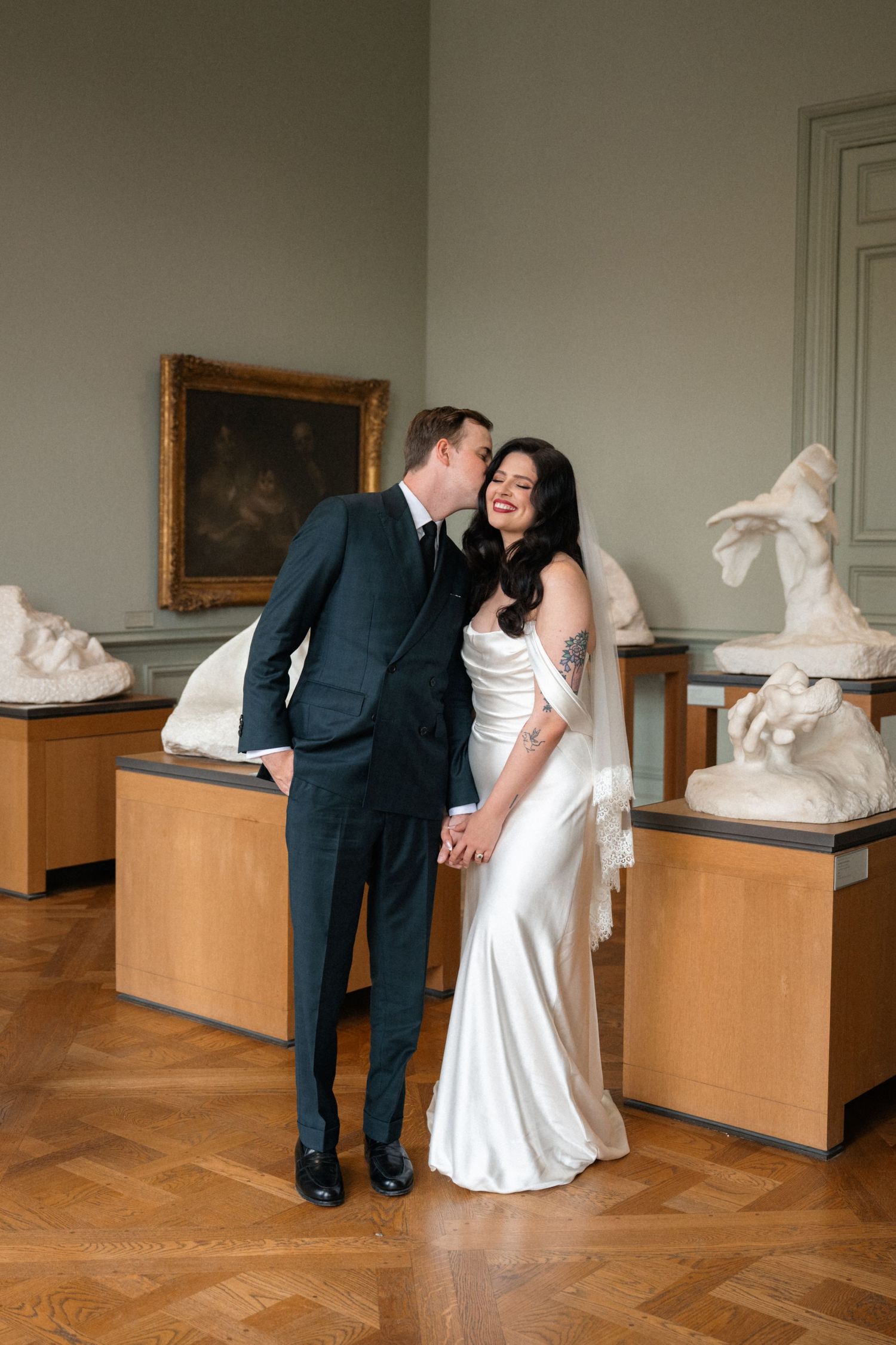 Groom whispers to laughing bride in white satin gown surrounded by classical sculptures in art museum.
