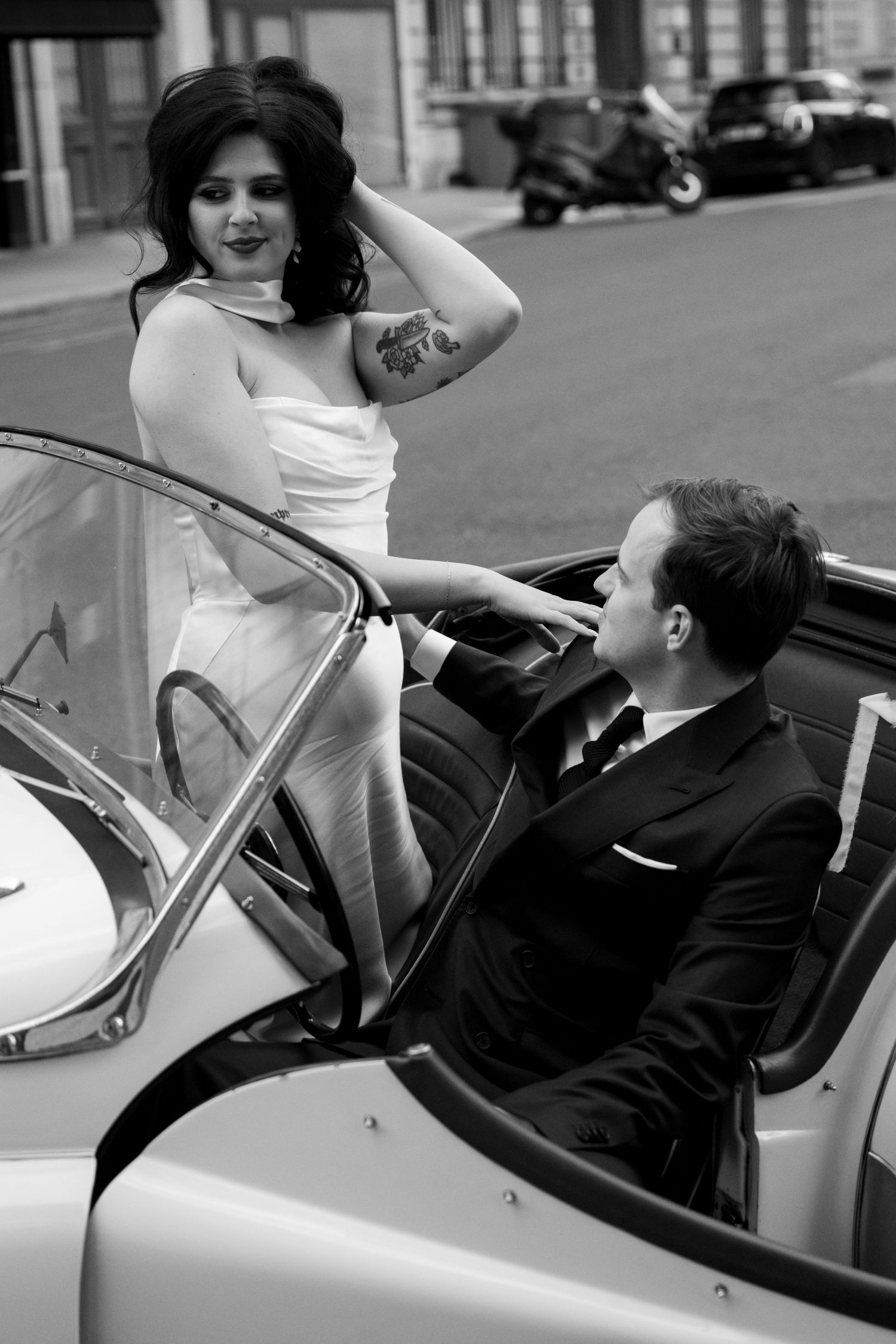 Black and white photo of bride posing with groom seated in vintage white convertible on Paris street.