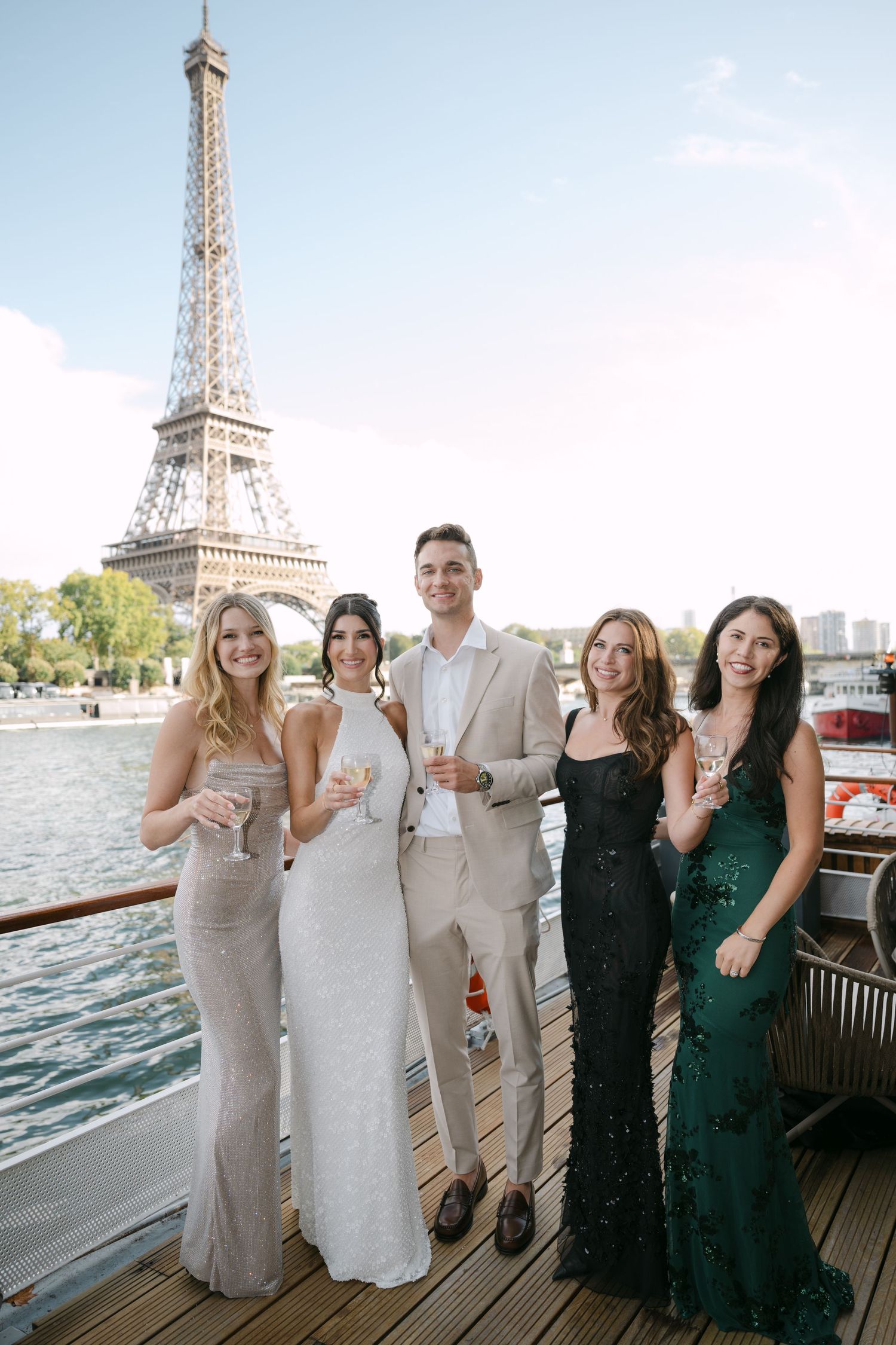 Five elegantly dressed wedding guests pose on a boat deck with the Eiffel Tower in Paris behind them.