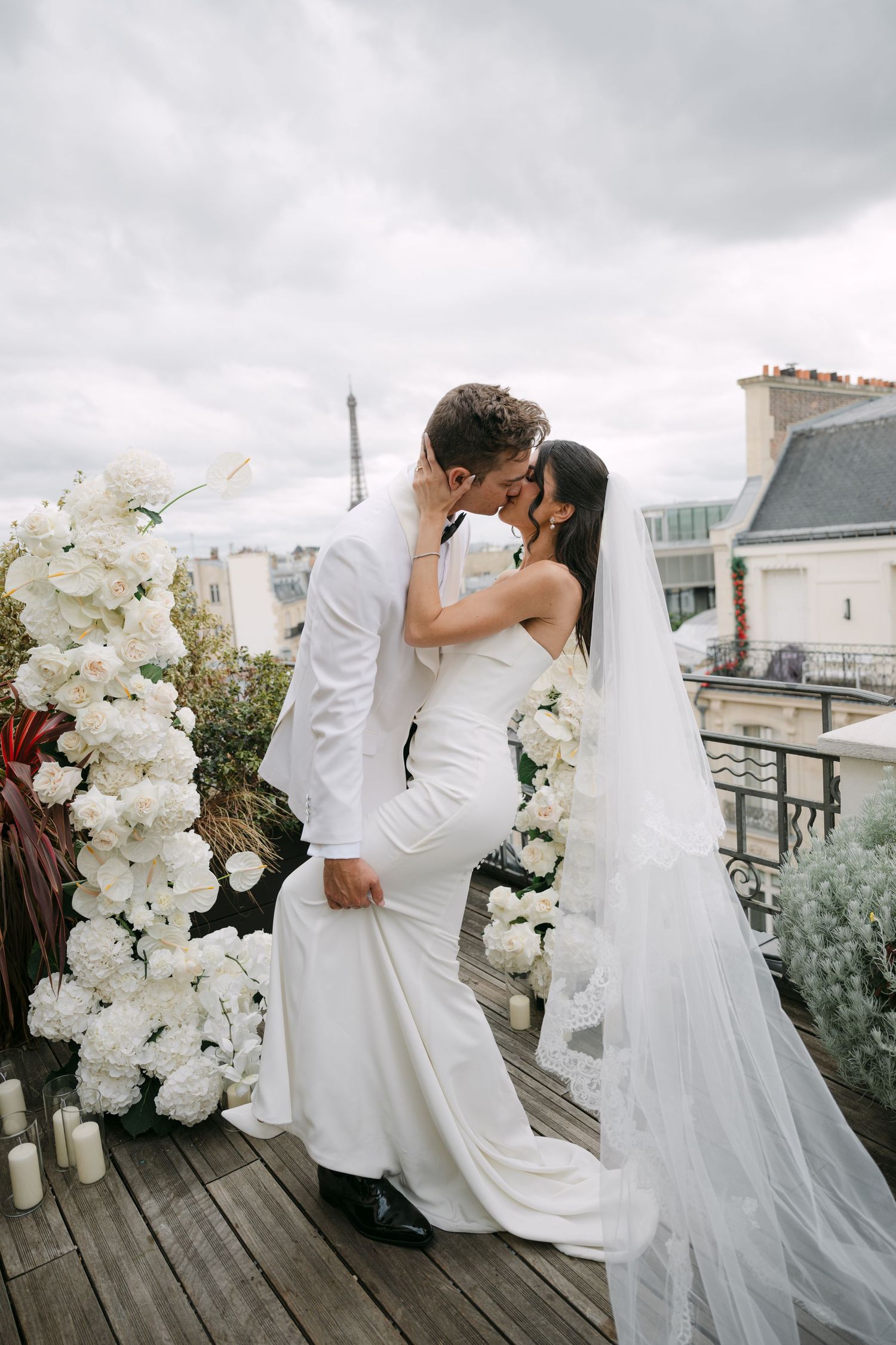 Bride and groom kissing beside white floral arch on Paris rooftop terrace.