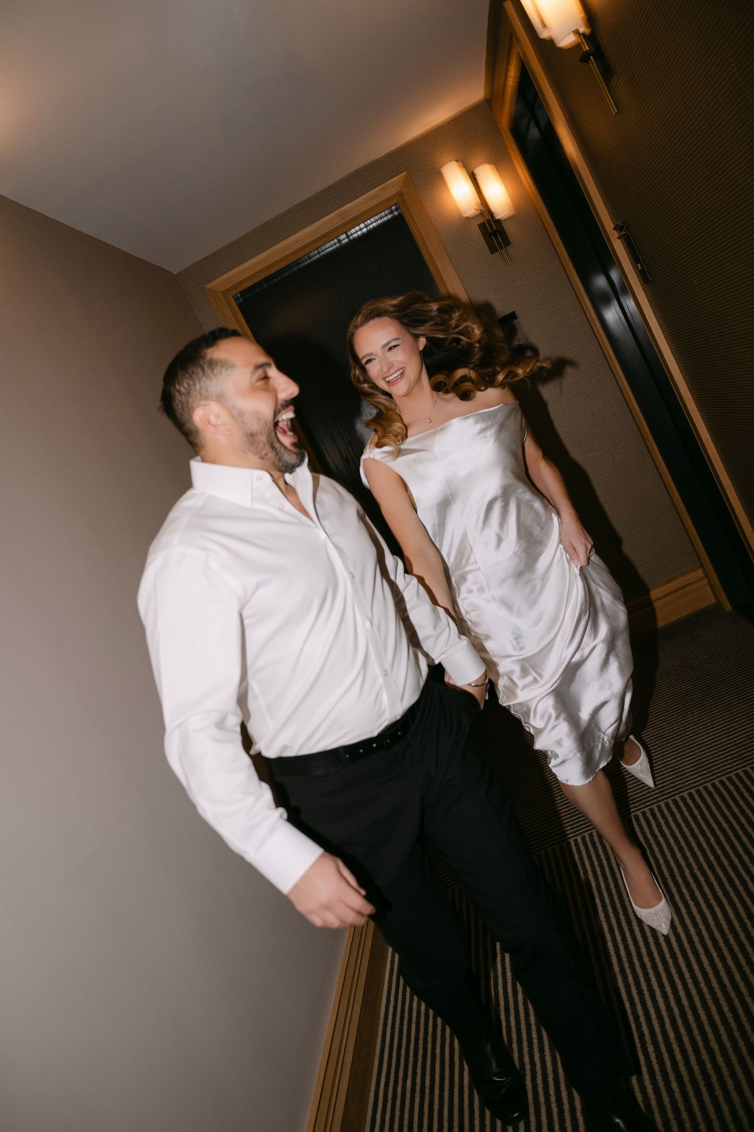 Laughing bride and groom walk down hotel hallway, she wears white dress while he wears white shirt and black pants.